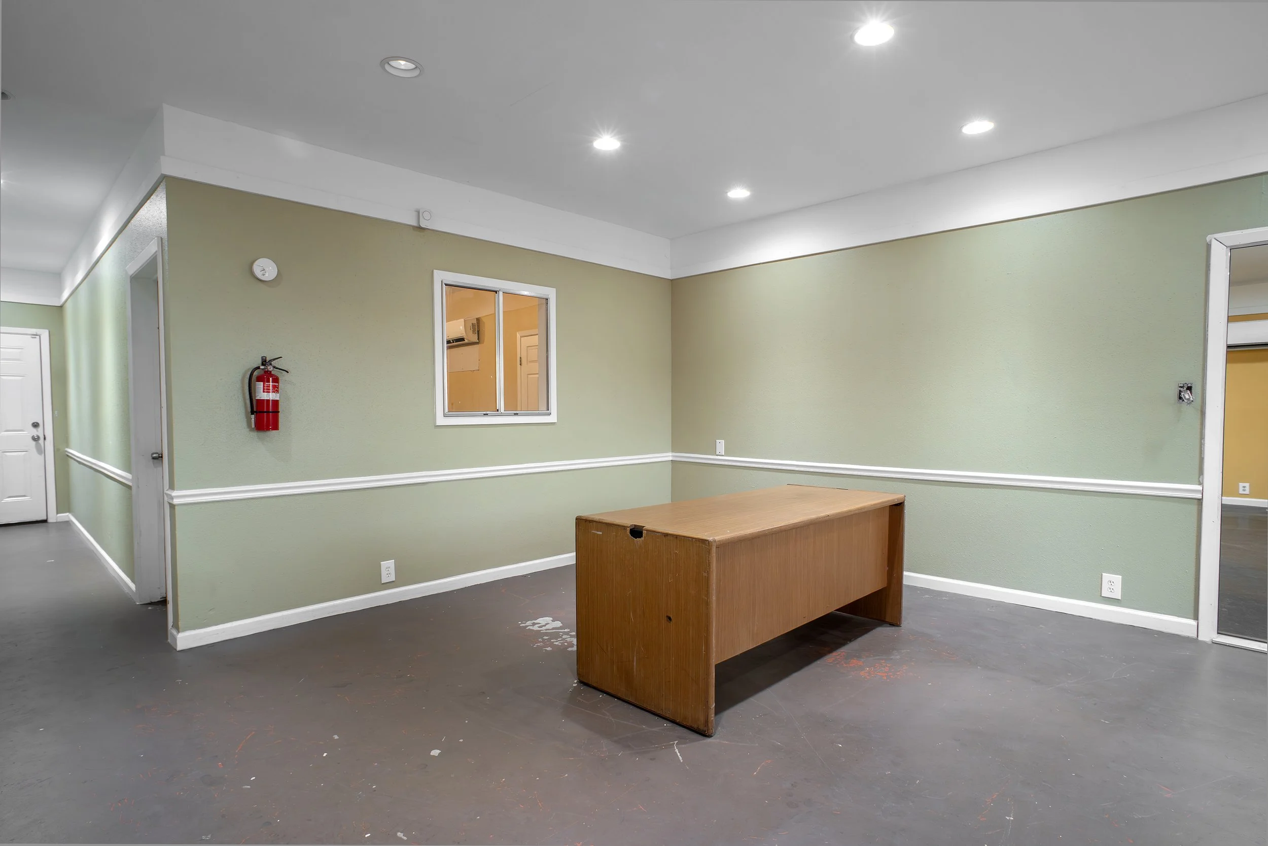An empty room with beige walls and a dark floor. There is a small wooden table and a window with a white frame. A fire extinguisher is mounted on the wall, and recessed lights are in the ceiling.