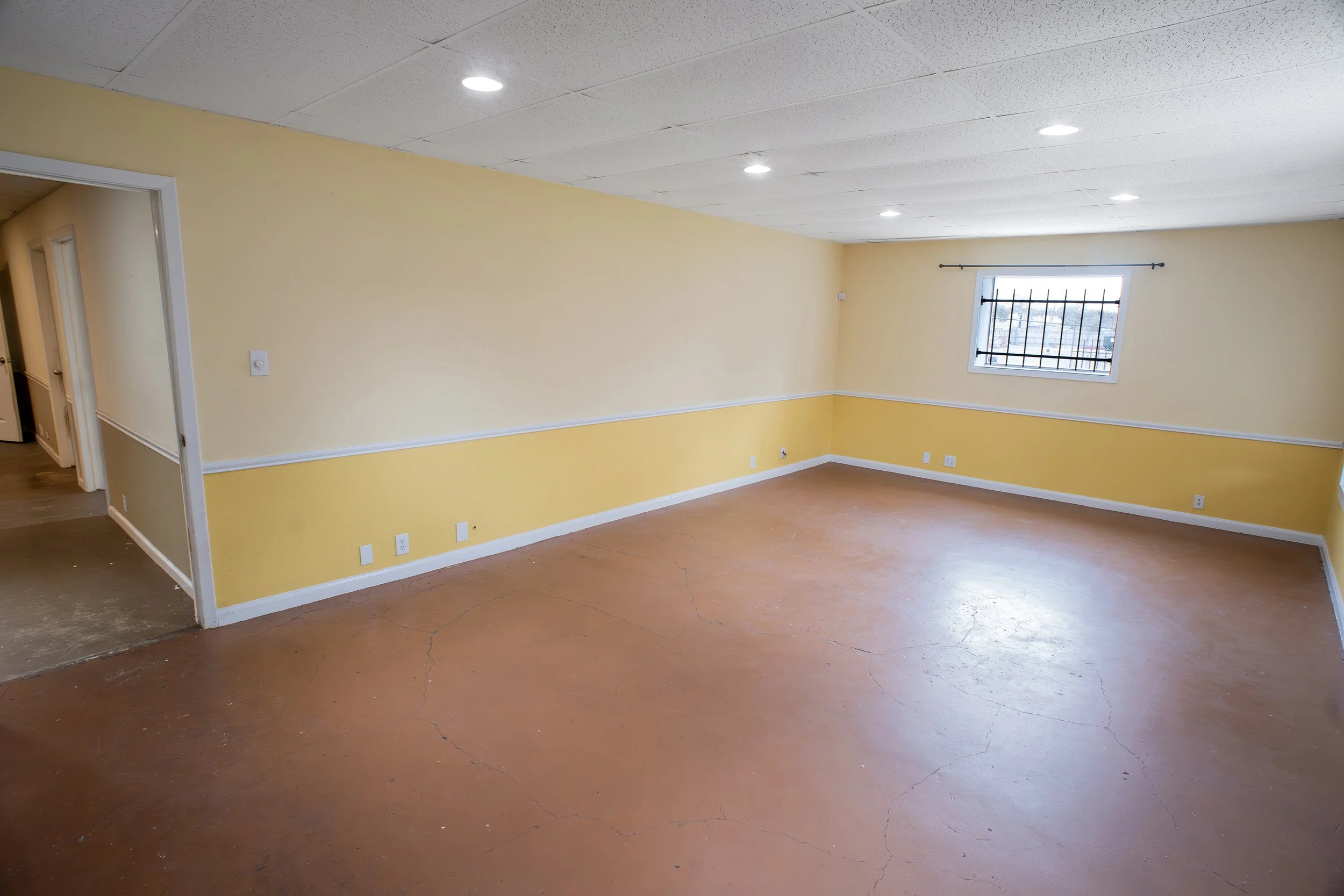 Empty room with yellow and white walls, a window with black bars, fluorescent ceiling lights, and a cracked brown floor.