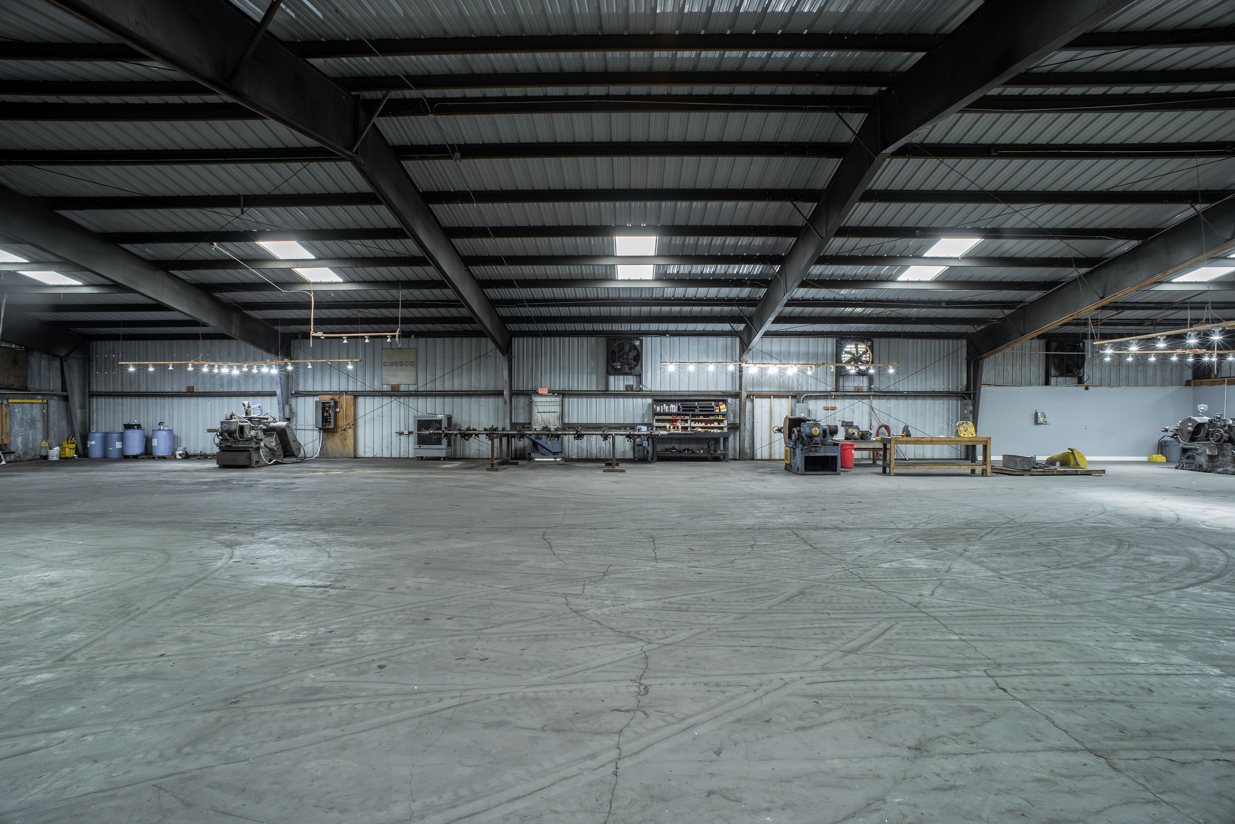 A large, empty industrial warehouse with a high, metal ceiling, bright lighting, and various machinery and storage shelves along the walls.