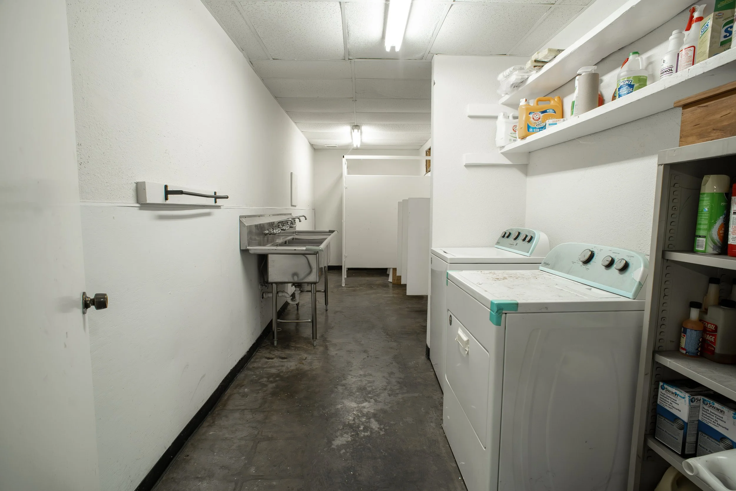 A narrow laundry room with white walls, a black baseboard, and a painted concrete floor. There are shelves with cleaning supplies, a washing machine, a utility sink, and fluorescent ceiling lights.