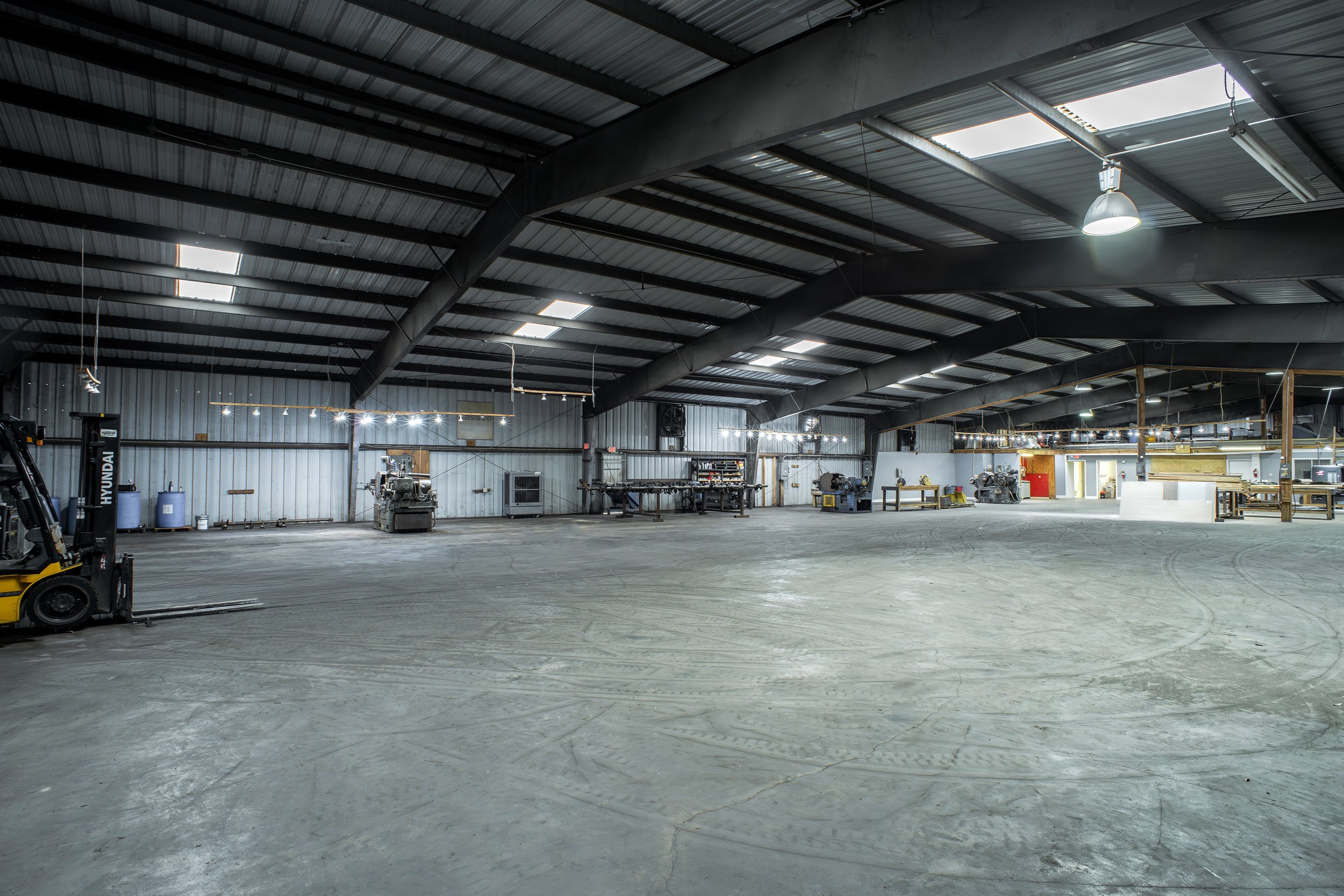 Interior of a large industrial warehouse with a metal roof, skylights, and various equipment and workstations, including a yellow forklift on the left.