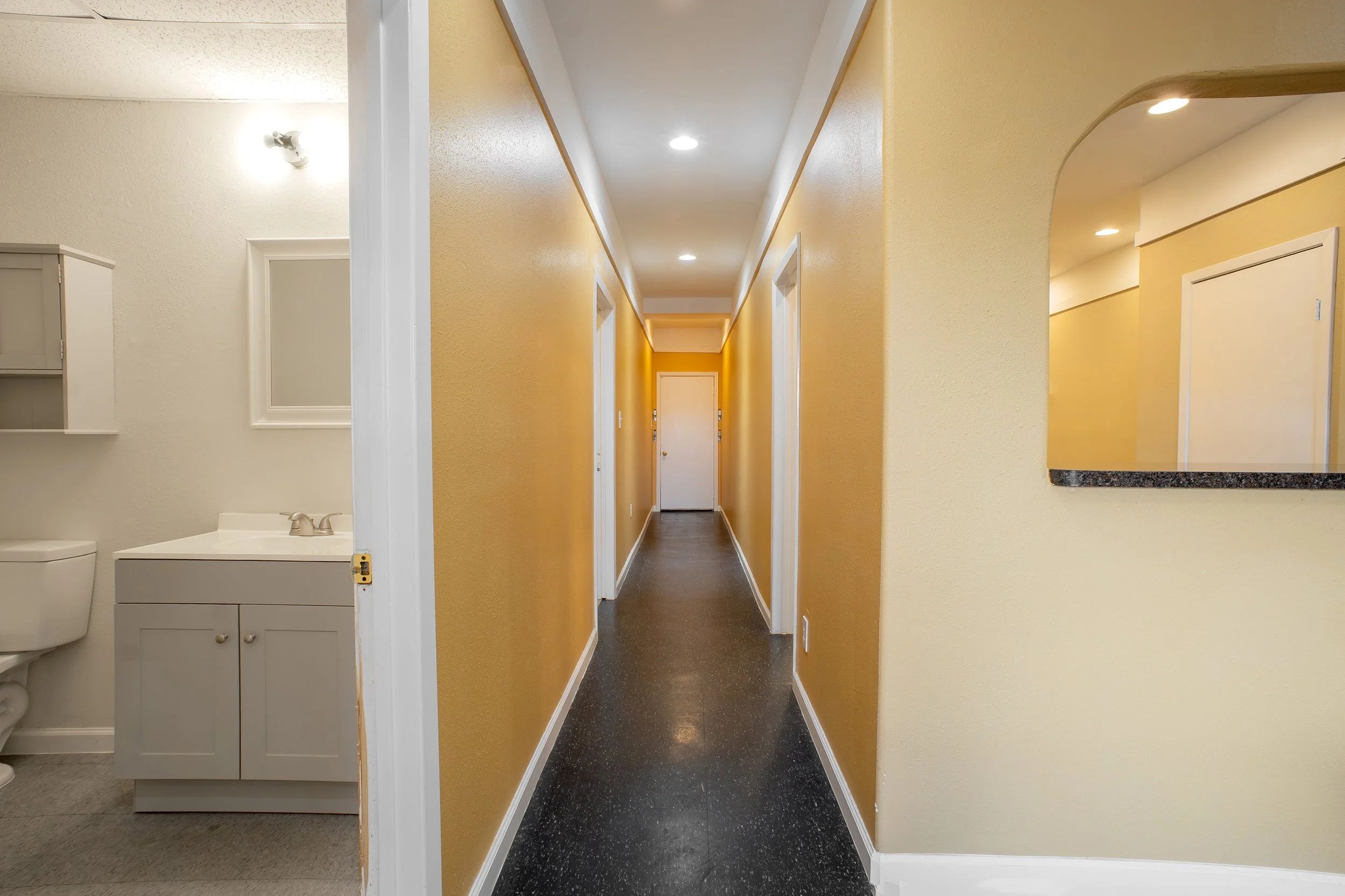 View of a narrow hallway with bright yellow walls, dark flooring, white ceiling, and doors at the end. To the left, part of a bathroom with a vanity, mirror, and toilet is visible. On the right, a framed mirror hangs on the wall.