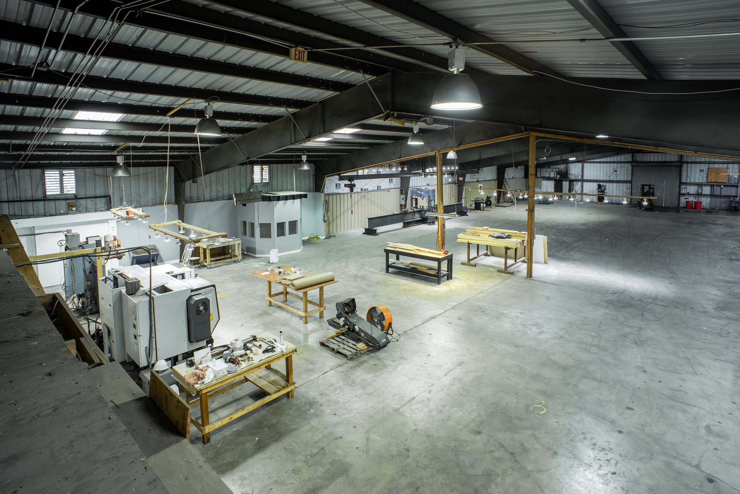 Interior of a spacious industrial warehouse or workshop with various equipment, workbenches, and tools. The high ceiling has metal beams and hanging lights, with some windows and an exit sign visible.