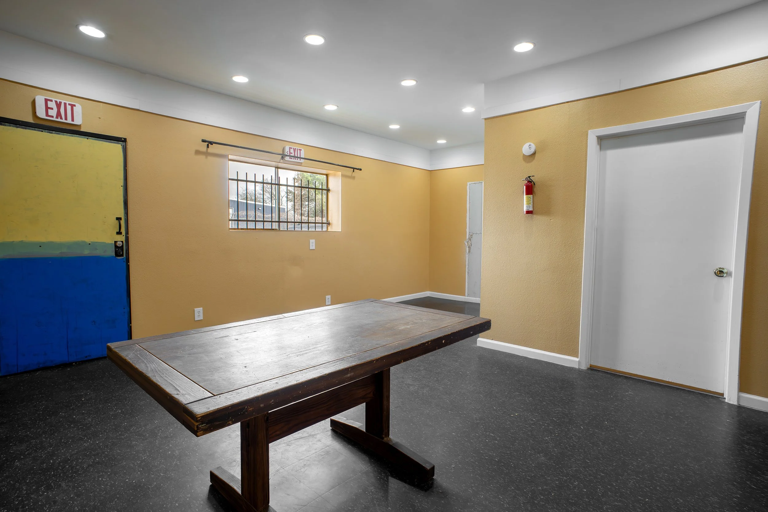 Interior of a room with yellow walls, a window with security bars, a fire extinguisher, and overhead lighting. There is a wooden table in the foreground.