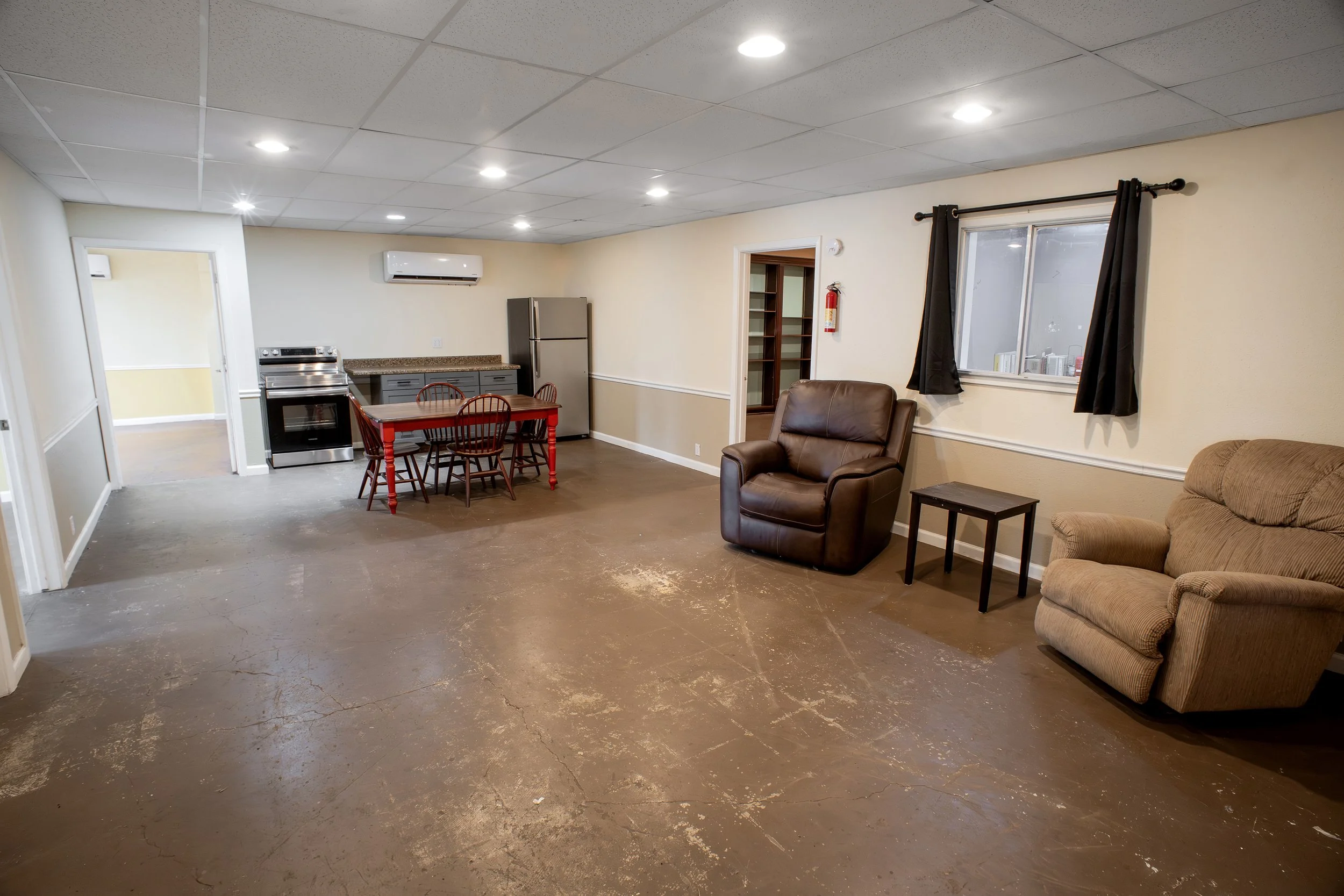 Living room with beige walls, ceiling lights, a window with black curtains, a small sofa, a recliner, a small side table, a kitchen area with a table and chairs, refrigerator, stove, and air conditioning unit.