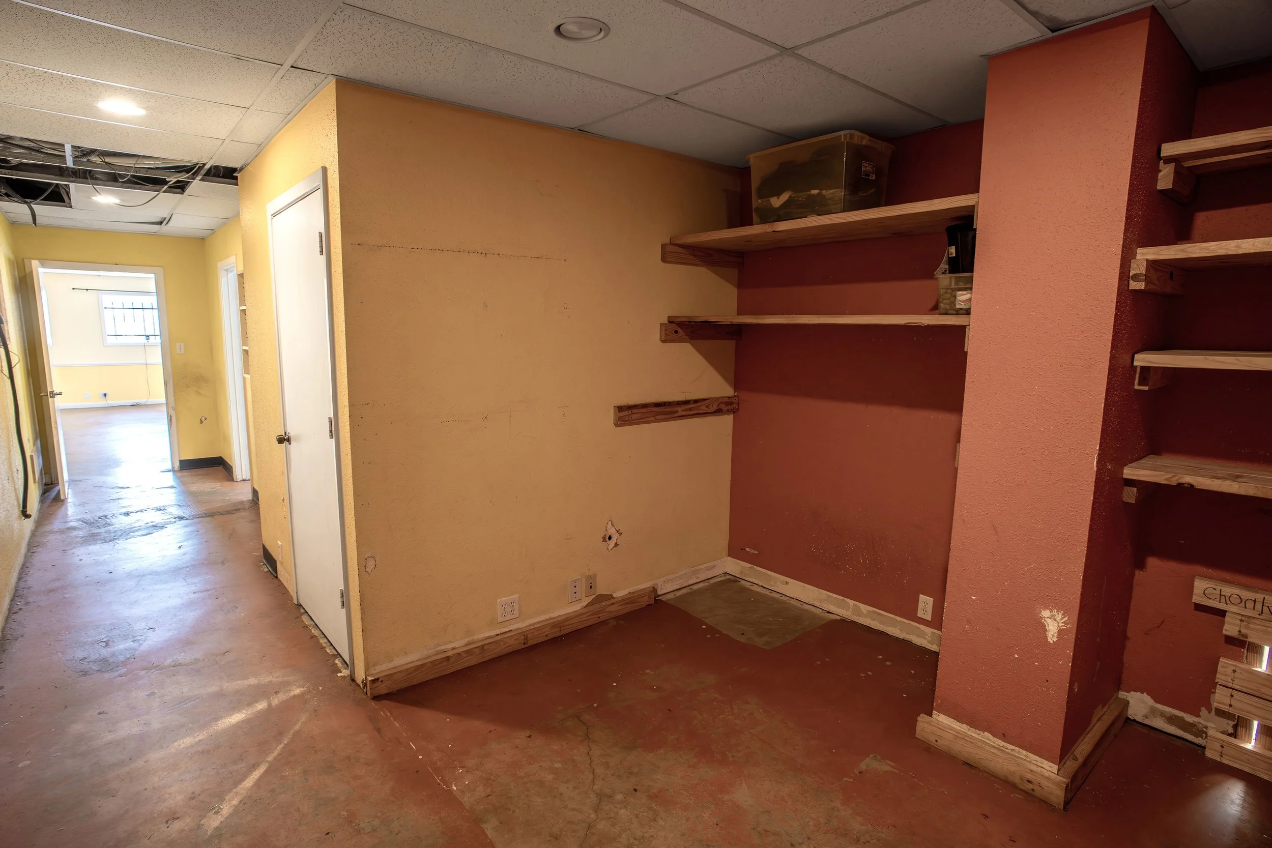 An interior room under renovation with unfinished walls and wooden shelves. The ceiling has visible wiring and missing ceiling tiles. The floor is concrete with paint stains and cracks. There are two windows and a door at the end of the hallway.