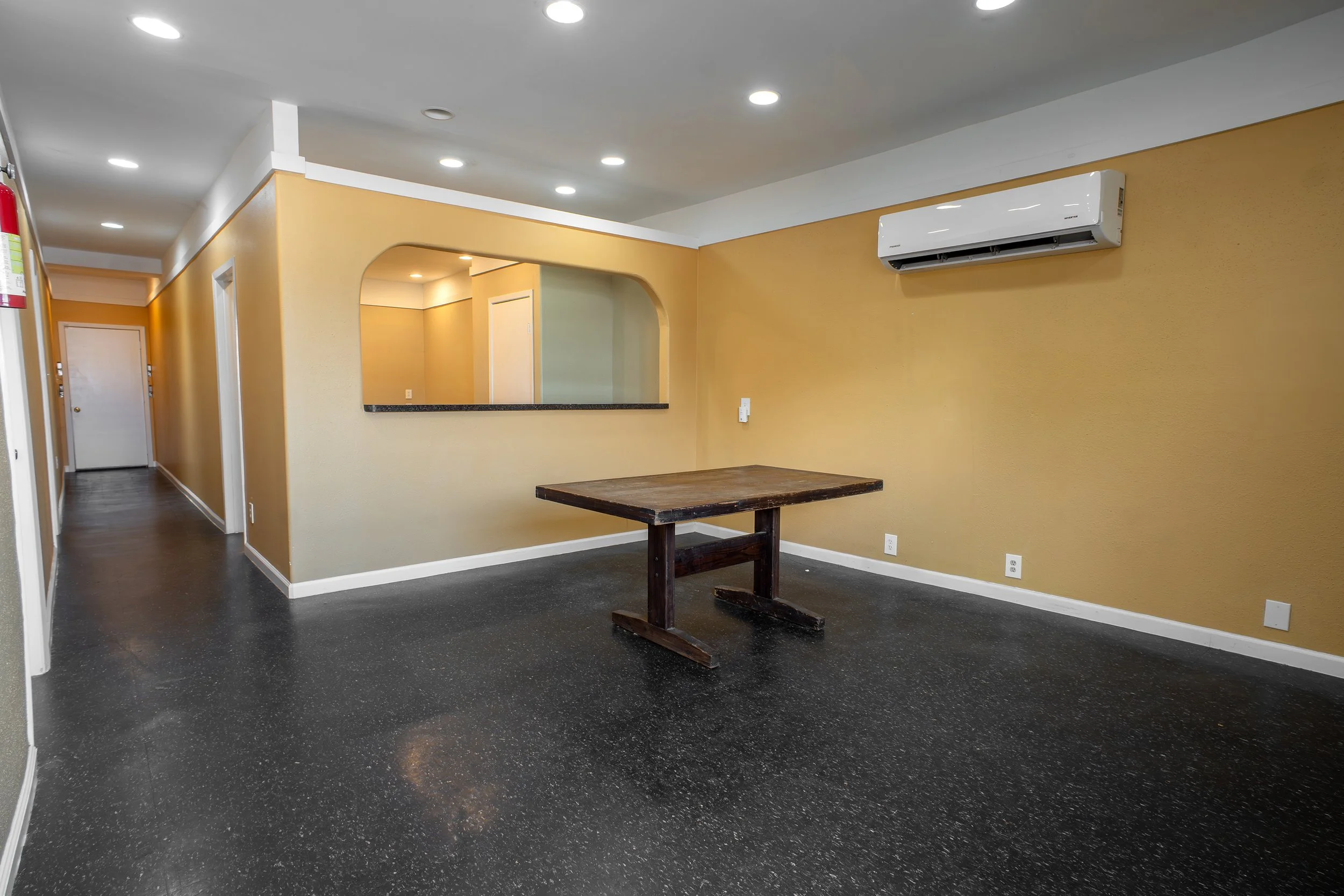 Interior of a room with yellow walls, a black speckled floor, a wooden table, and an air conditioning unit on the wall. There is a mirror on the wall reflecting part of the room, and a white door in the background.