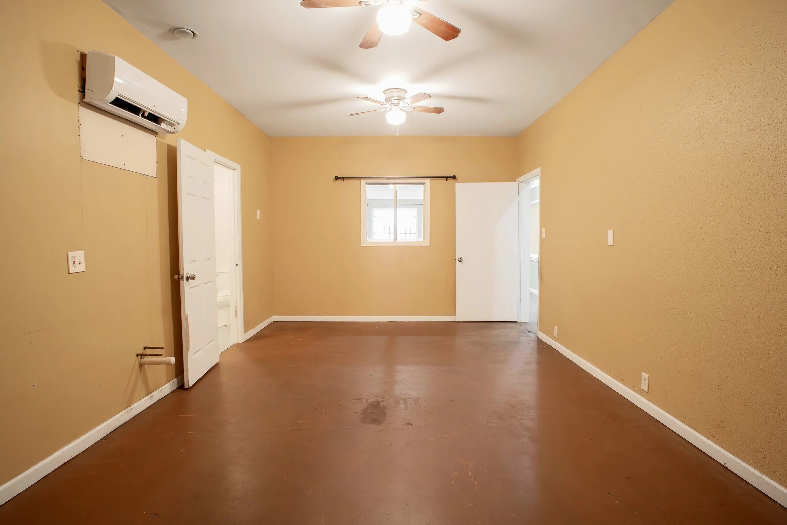 Empty room with yellow walls, brown hardwood floor, ceiling fans with wood blades, a window, and an air conditioning unit mounted on the wall.
