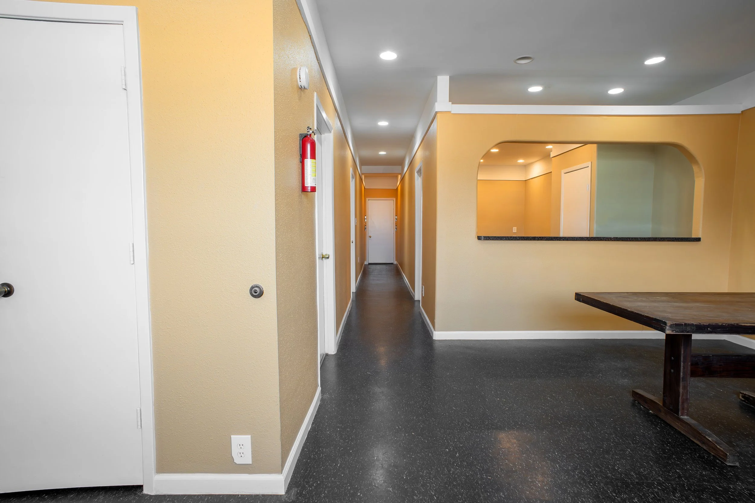 Image of an indoor hallway with yellow walls and a dark speckled floor. There is a fire extinguisher mounted on the wall, and doorways leading to other rooms. An opening in the wall reveals a view of a room with a dark countertop.