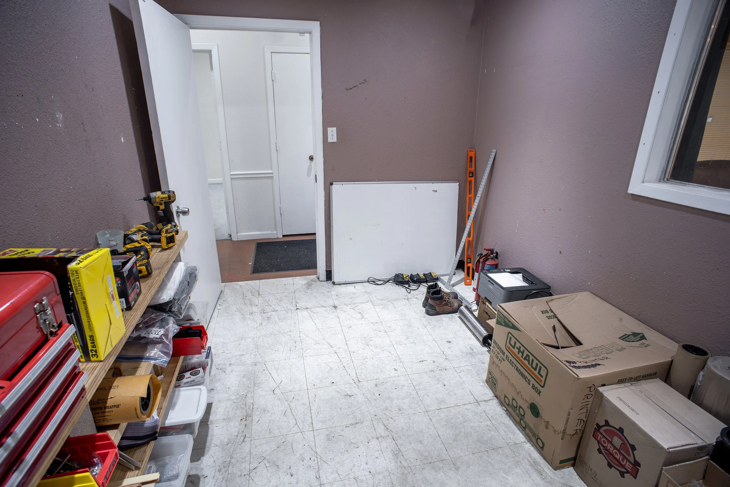 A cluttered workshop or storage room with tools, boxes, and construction materials. There is a door slightly open leading to another room, and various items like a level, power tools, shoes, and storage boxes are visible.