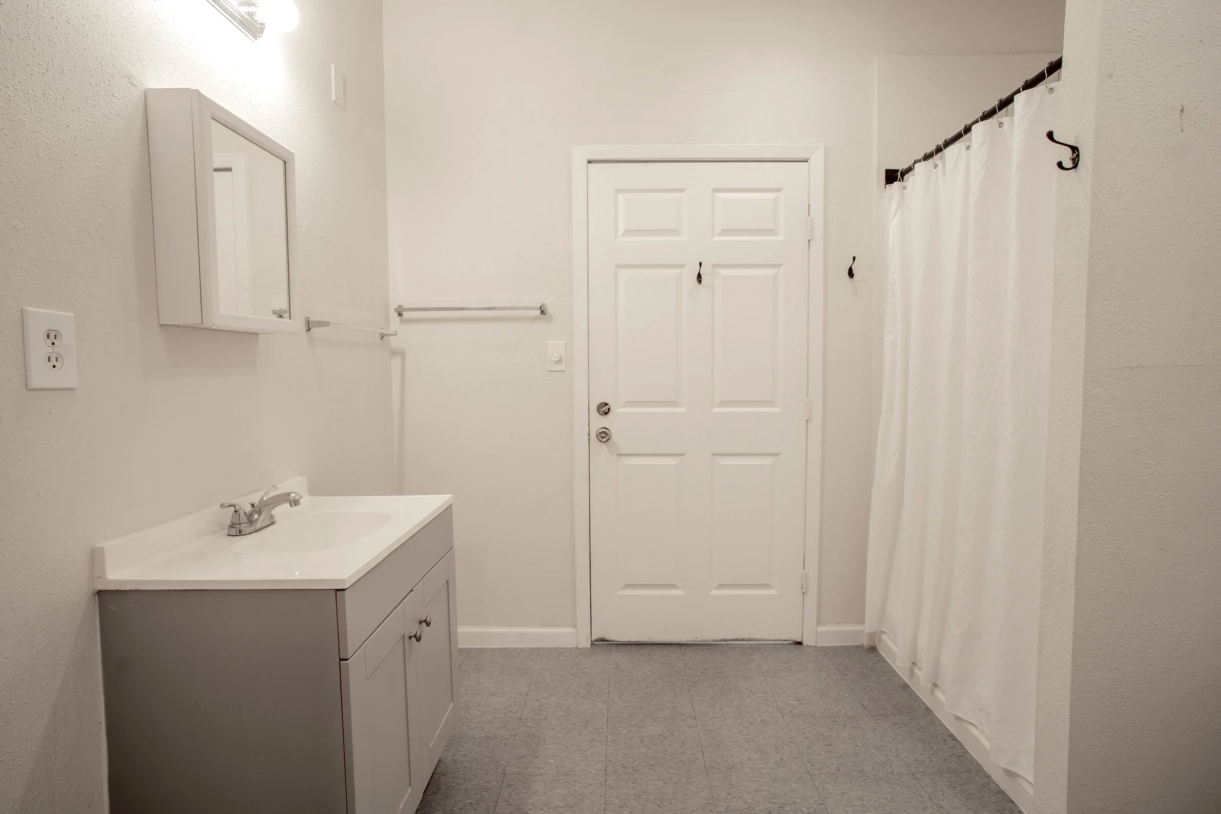A small bathroom with a white door, a white vanity with a sink, a mirror cabinet, a towel bar, and a shower with white curtains.