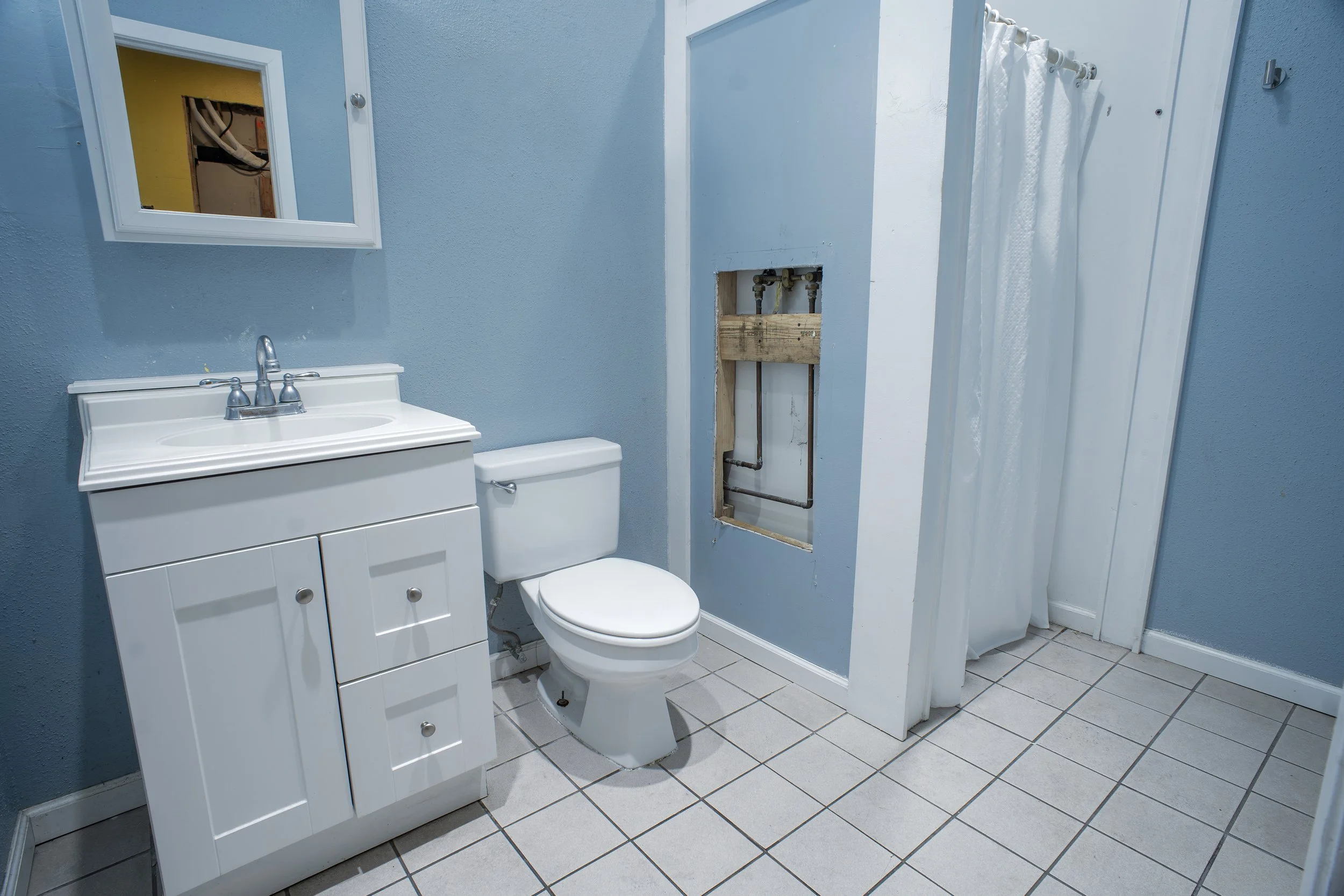 A small bathroom with light blue walls, a white vanity with a metal faucet, a white toilet, a mirror above the vanity, and a partially removed shower stall with exposed pipes and a white shower curtain.