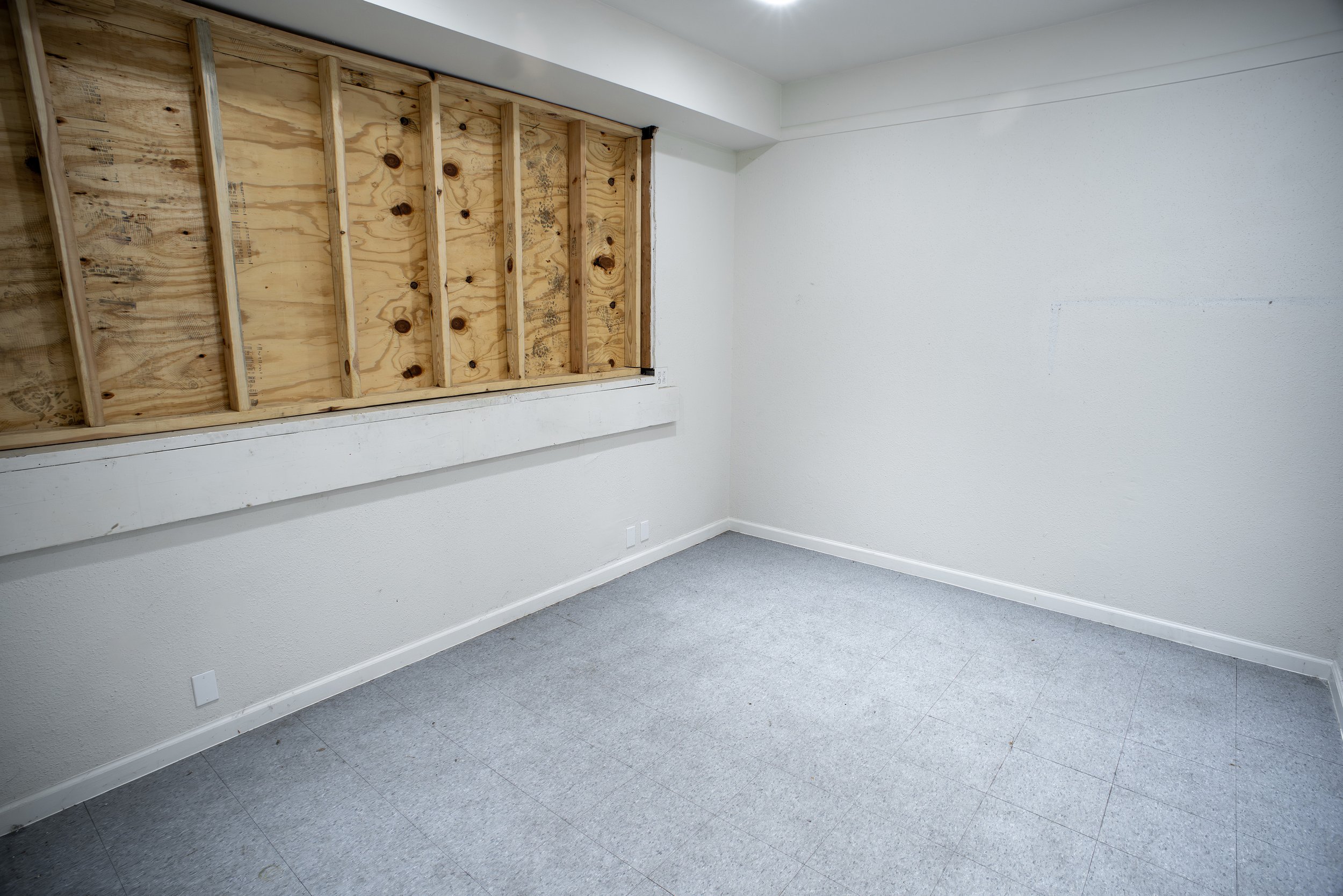 Empty room with white walls, gray tiled floor, and a large window opening with exposed wooden framing, no glass or window installed.