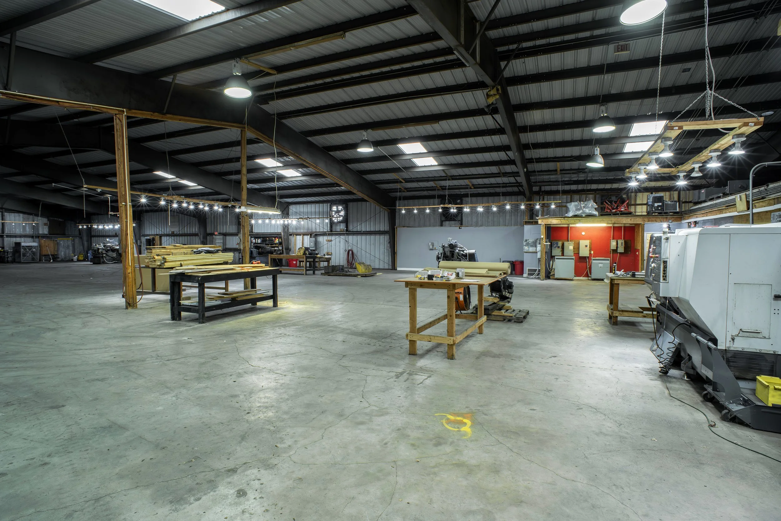 Interior of a large industrial workshop with workbenches, equipment, overhead lighting, and a high metal ceiling.