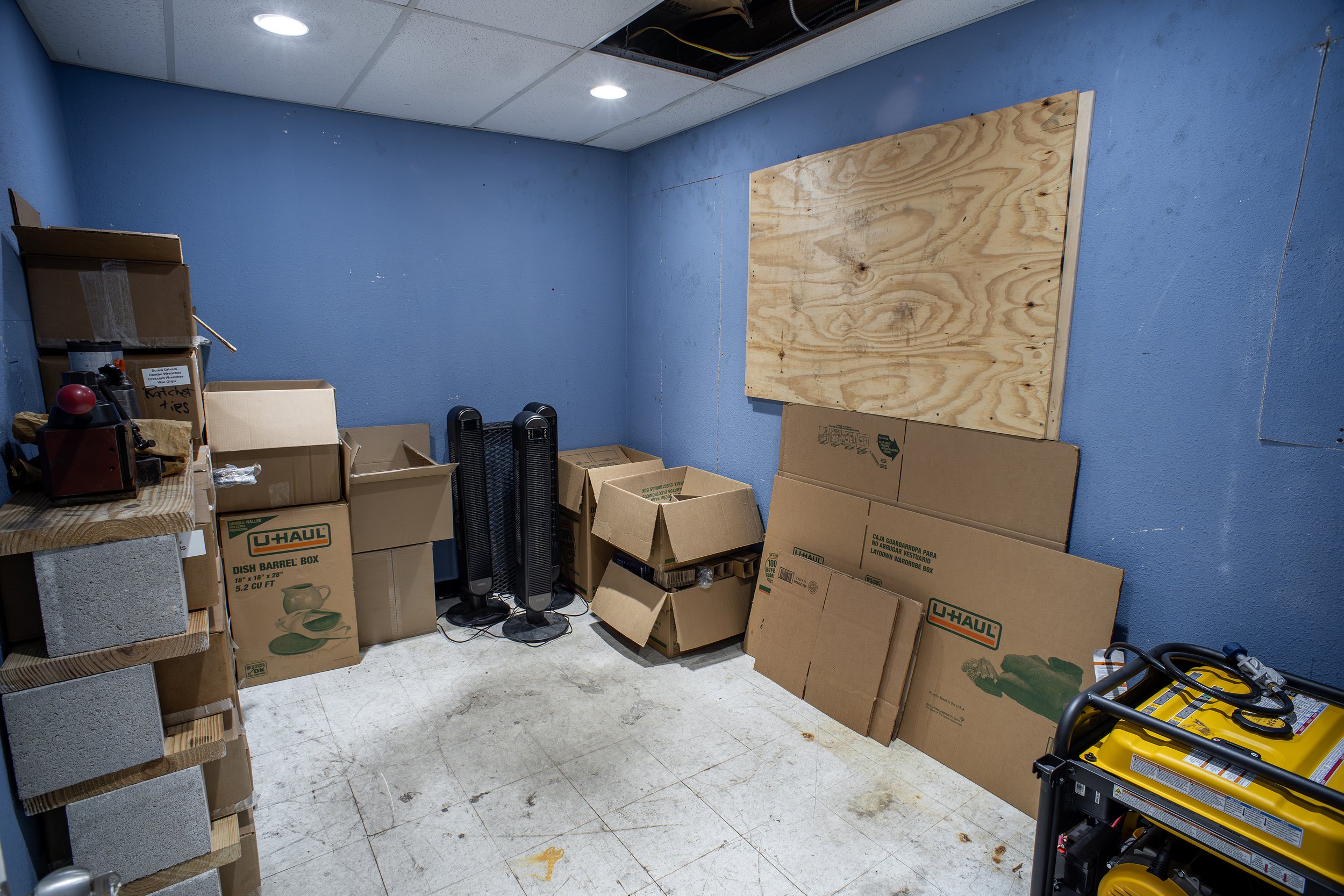 A storage room with blue walls and a white tiled floor, filled with cardboard boxes, some labeled 'U-Haul'. There is a large plywood board leaning against the wall, and some small appliances and tools are on shelves and the floor, including space hea