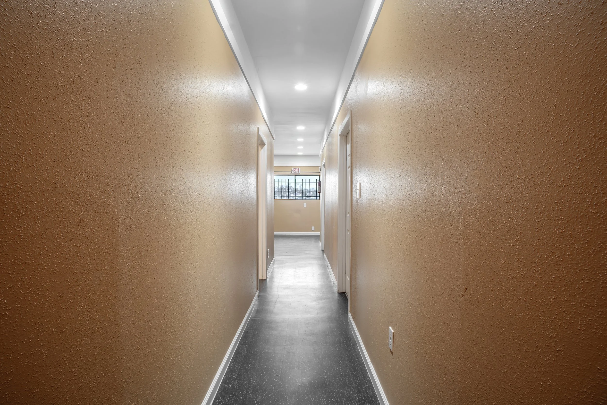 Empty hallway with beige textured walls, white ceiling with built-in lights, and gray flooring. Doors on the sides and a window at the end of the hallway.