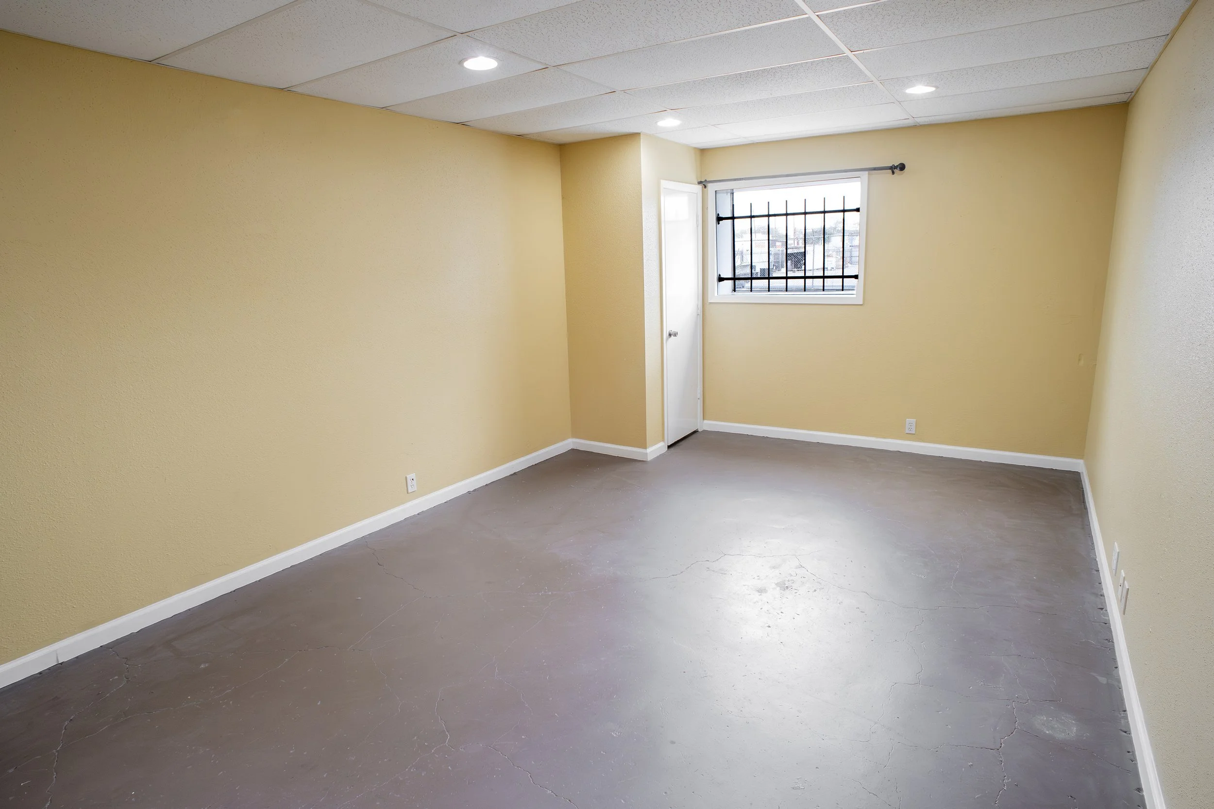 Empty room with yellow walls, a window with bars, a closed door, and a gray floor with visible cracks.