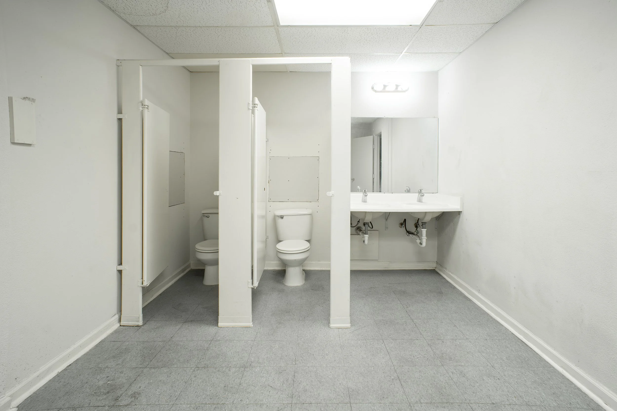 A public men's restroom with two toilet stalls and sinks, all painted white, with a mirror above the sinks and a fluorescent light on the ceiling.