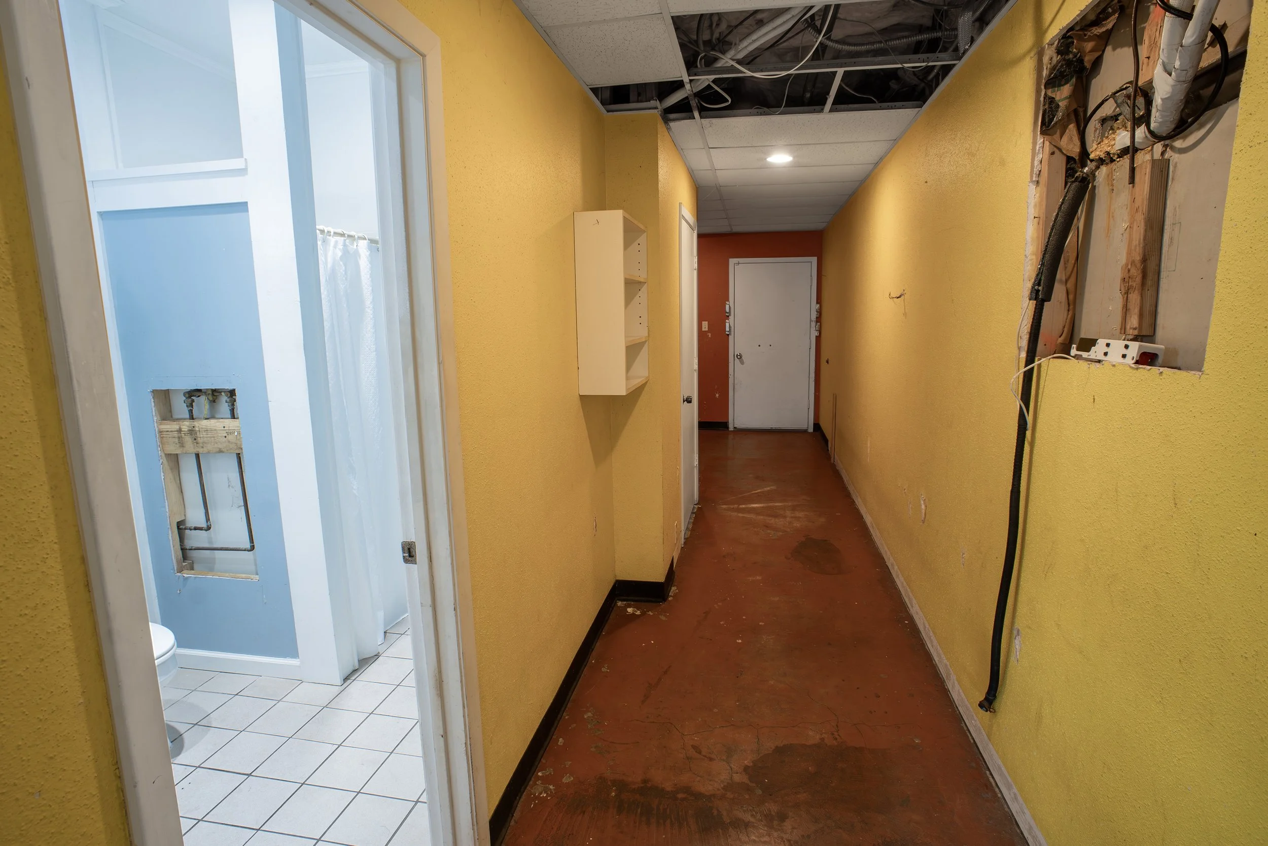 A hallway with yellow walls and a damaged ceiling with exposed wires. On the right side, a hole in the wall reveals electrical wiring and plumbing. The floor is reddish-brown, and there is a white cabinet on the wall. At the end of the hallway, a whi