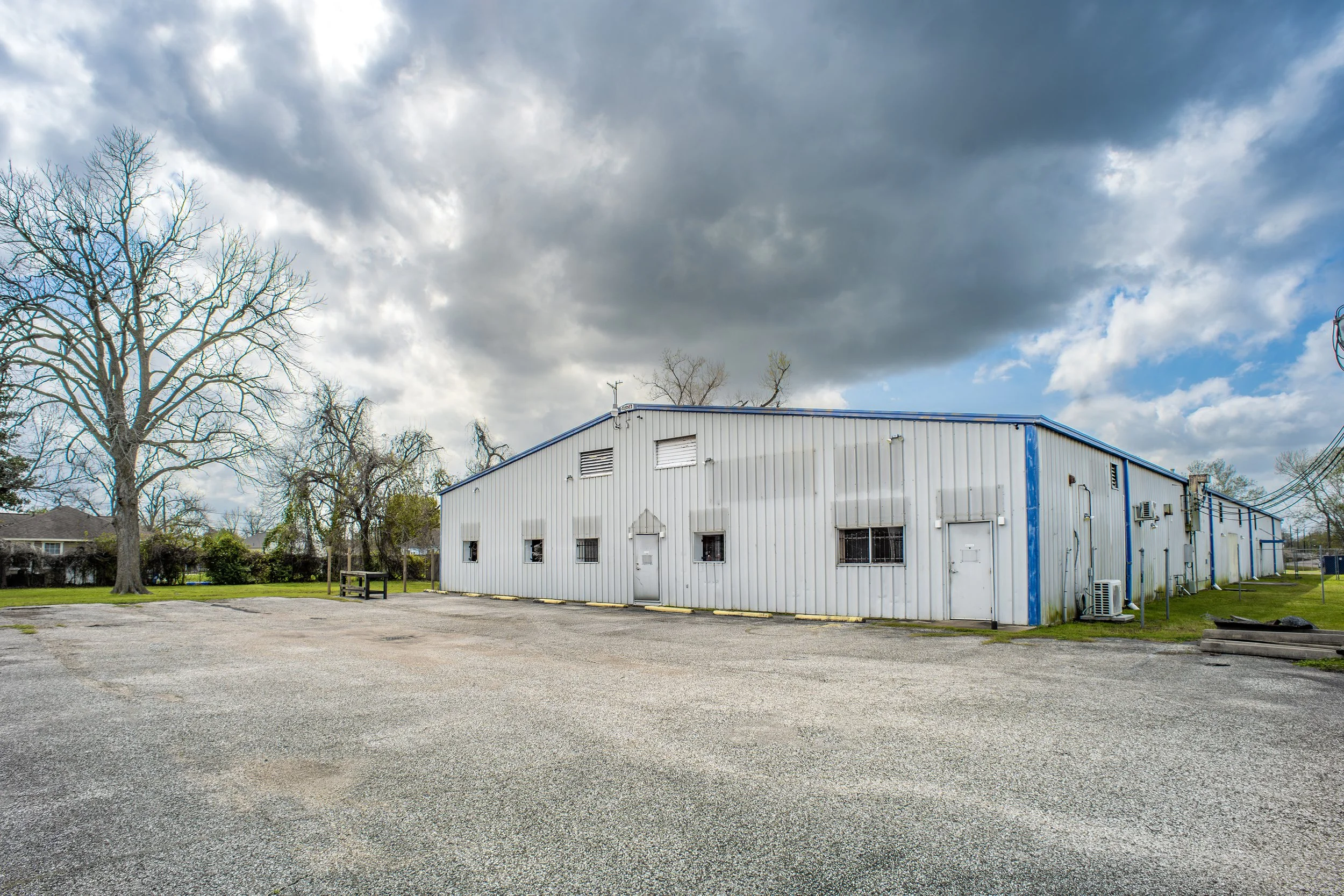 An industrial building with a metal exterior, blue accents, and small windows under a cloudy sky. In front, there is a paved lot with some patches of grass and a few trees nearby.