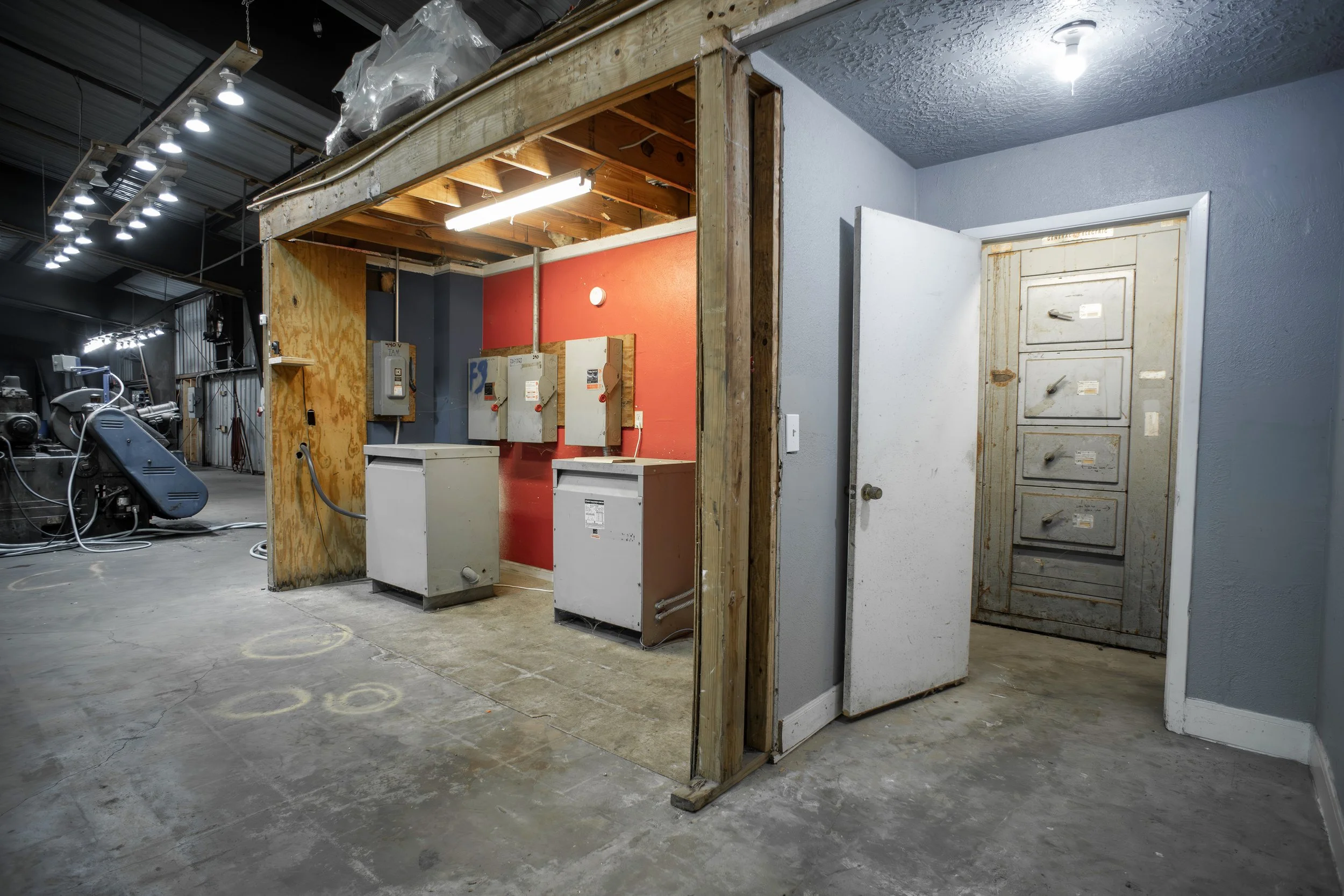 View of an unfinished wall in a garage or workshop, with visible electrical panels, tools, and industrial equipment, and exposed wooden framing and ceiling beams.