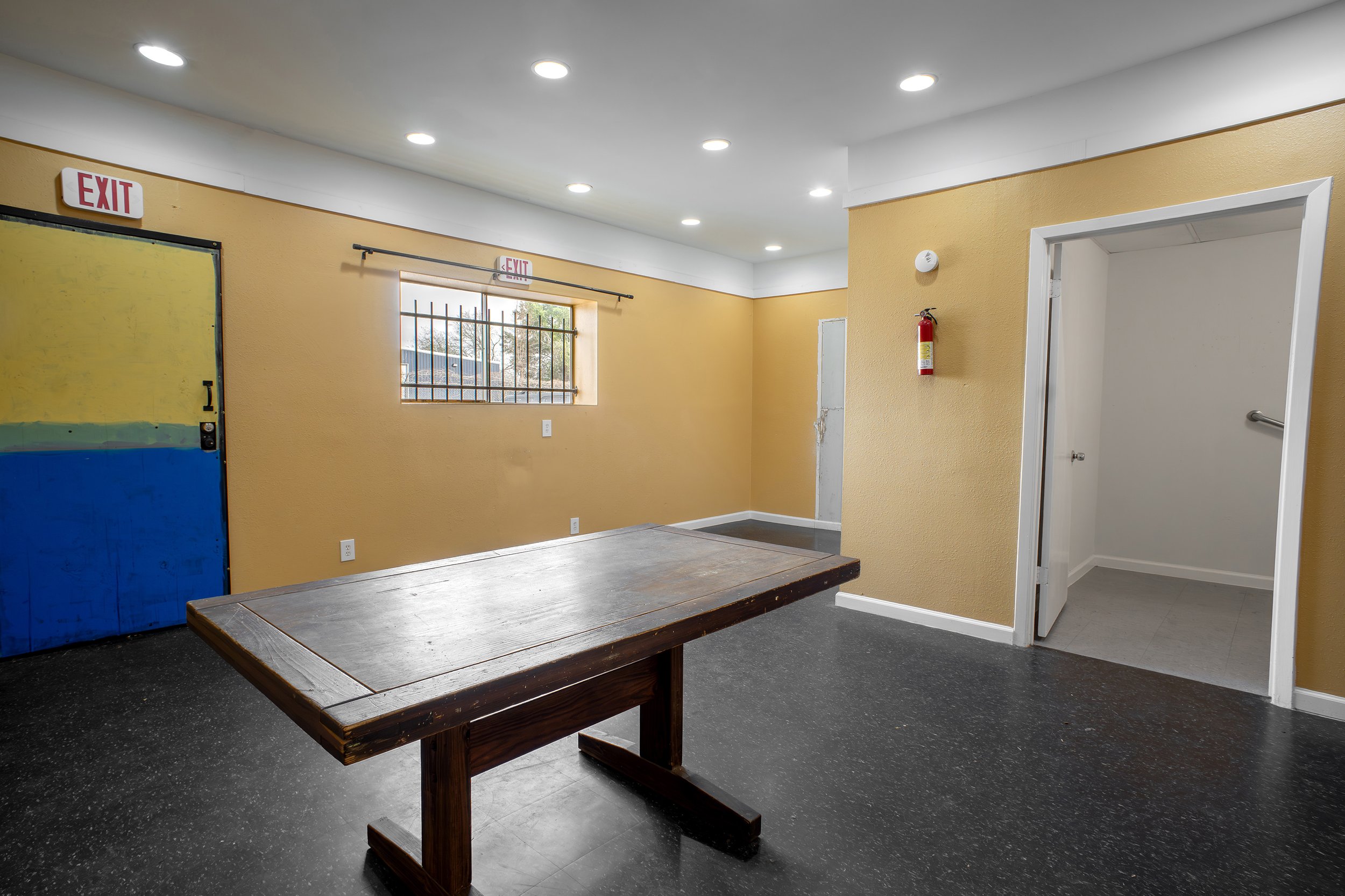 Interior view of a room with yellow walls, a window with bars, a fire extinguisher, a doorway, a wooden table, and ceiling lights.