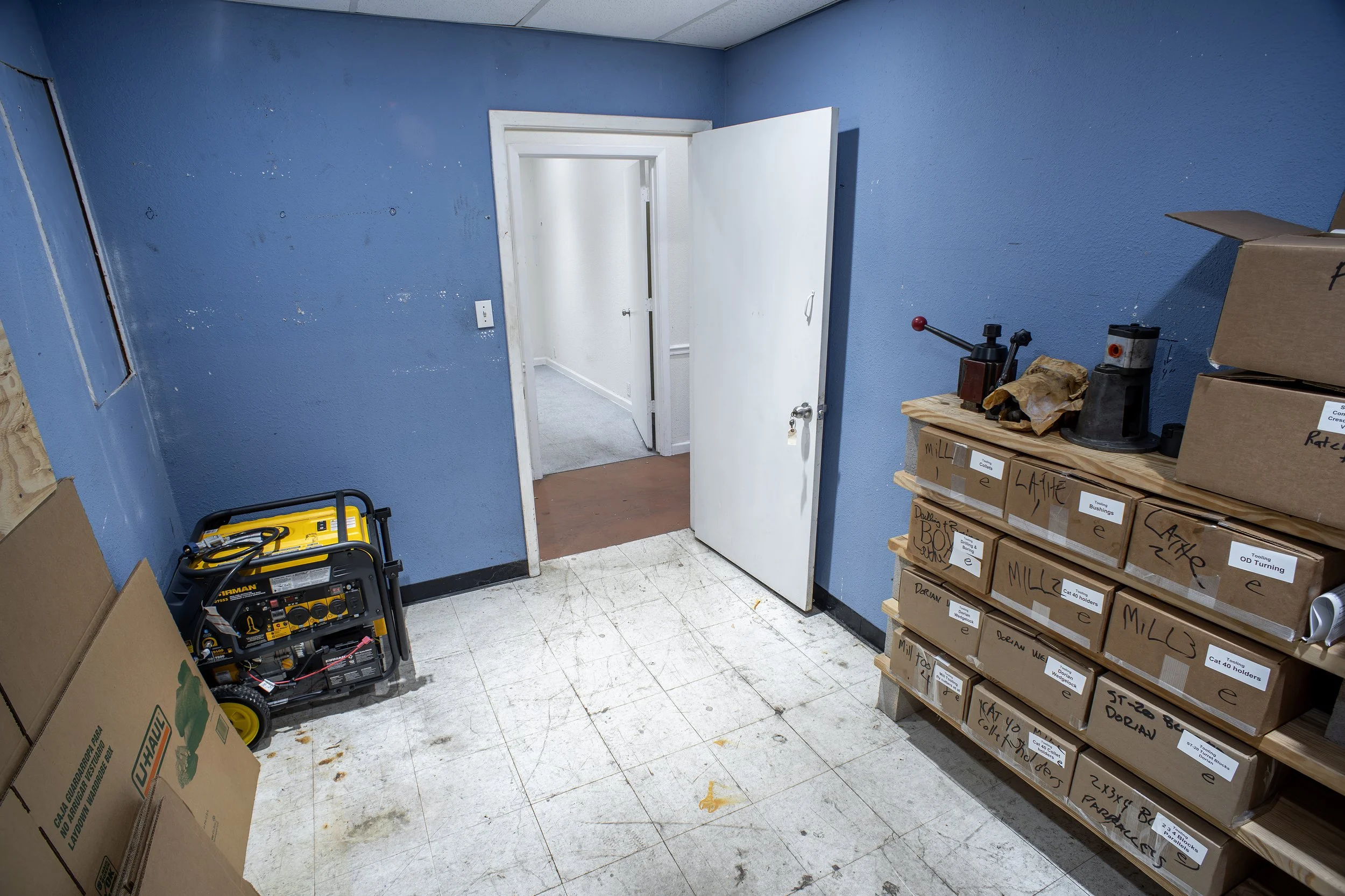 A small room with blue walls, white ceiling, and tiled floor, containing cardboard boxes, shelves with labeled boxes, and a yellow portable generator.