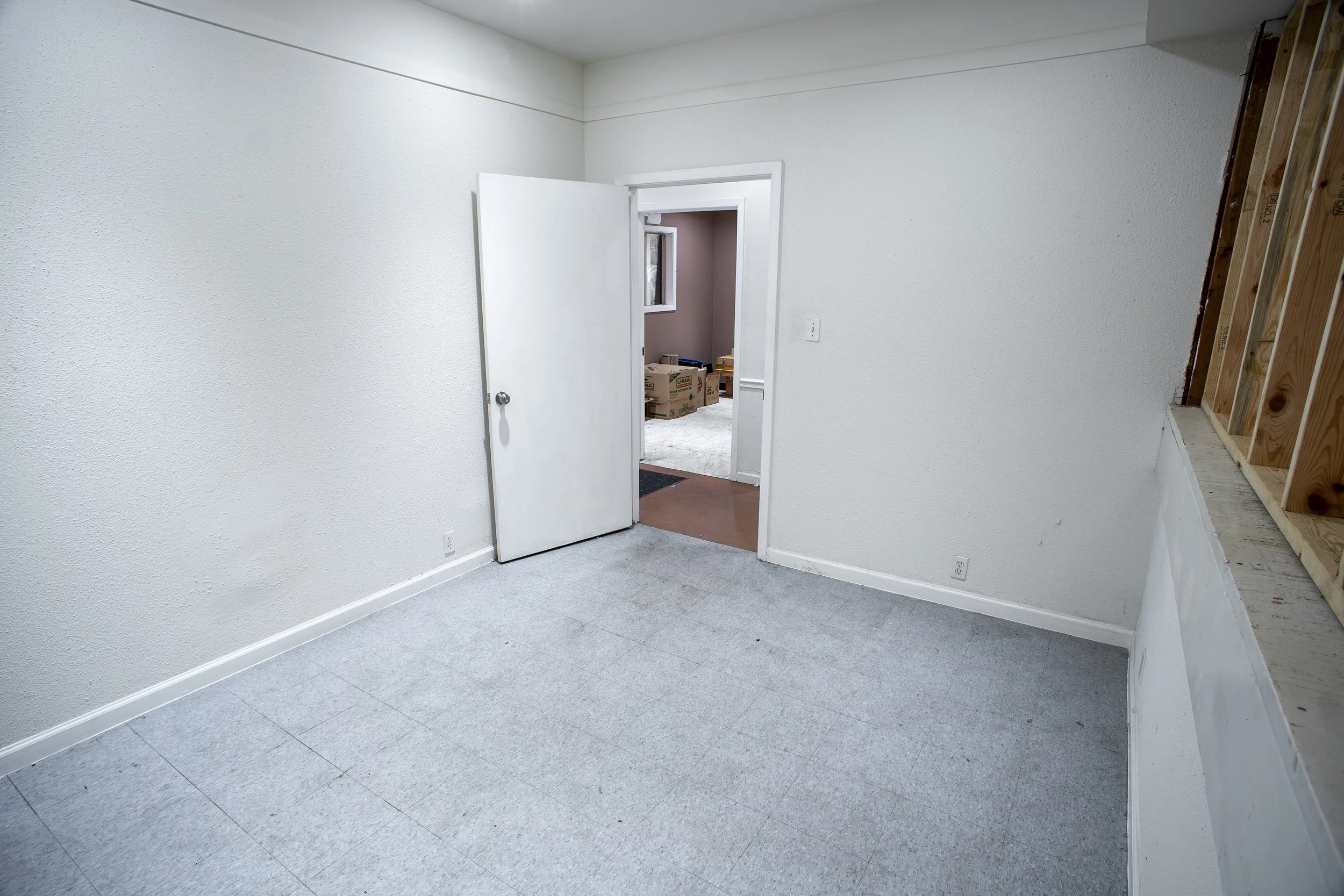 An empty room under construction with white textured walls, a door open to another room with boxes, and a partially built wall on the right with exposed wooden framing.