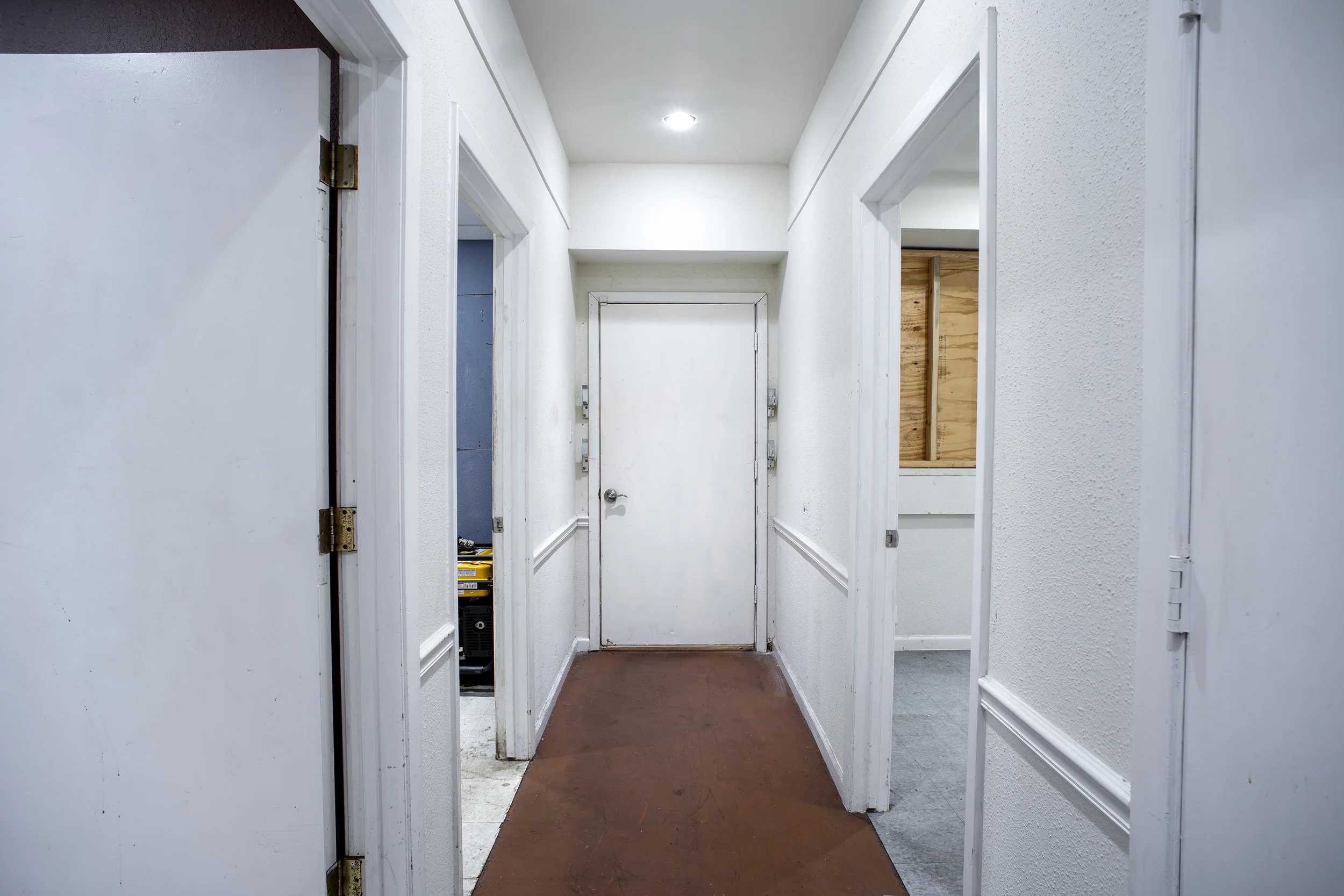 A narrow hallway with white walls, doorways on both sides, and a brown floor. The hallway has a ceiling light,