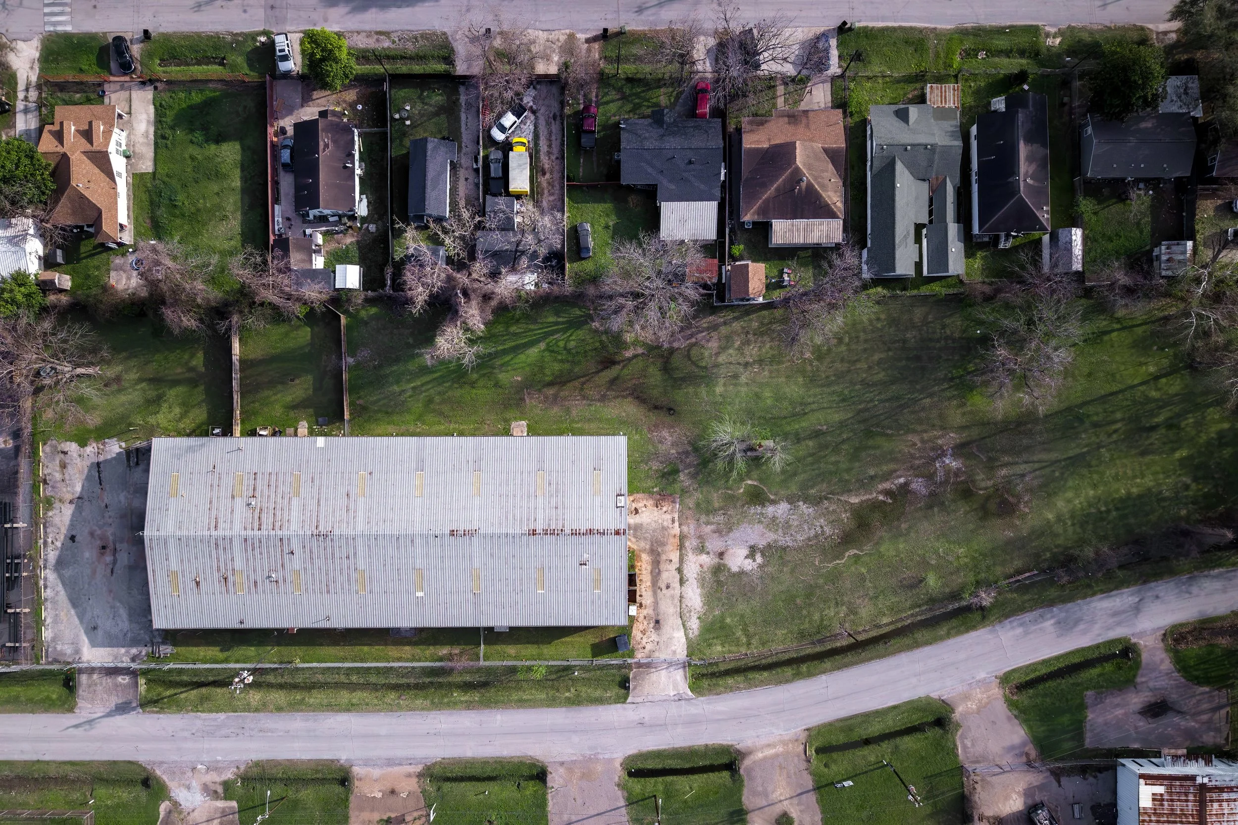 An aerial view of a suburban neighborhood showing houses, cars parked in driveways, trees, and yards, with a street running along the top of the image.