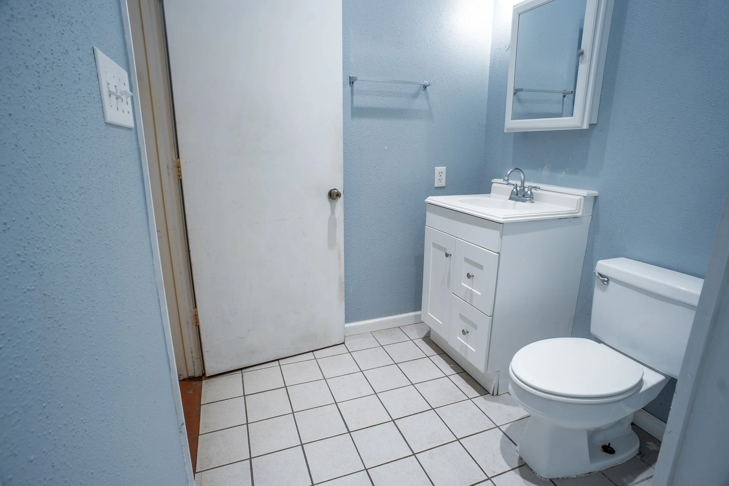 A small bathroom with light blue textured walls, a white toilet, a white vanity with a sink, a mirror cabinet, and a towel rack. The floor is tiled with light-colored square tiles.