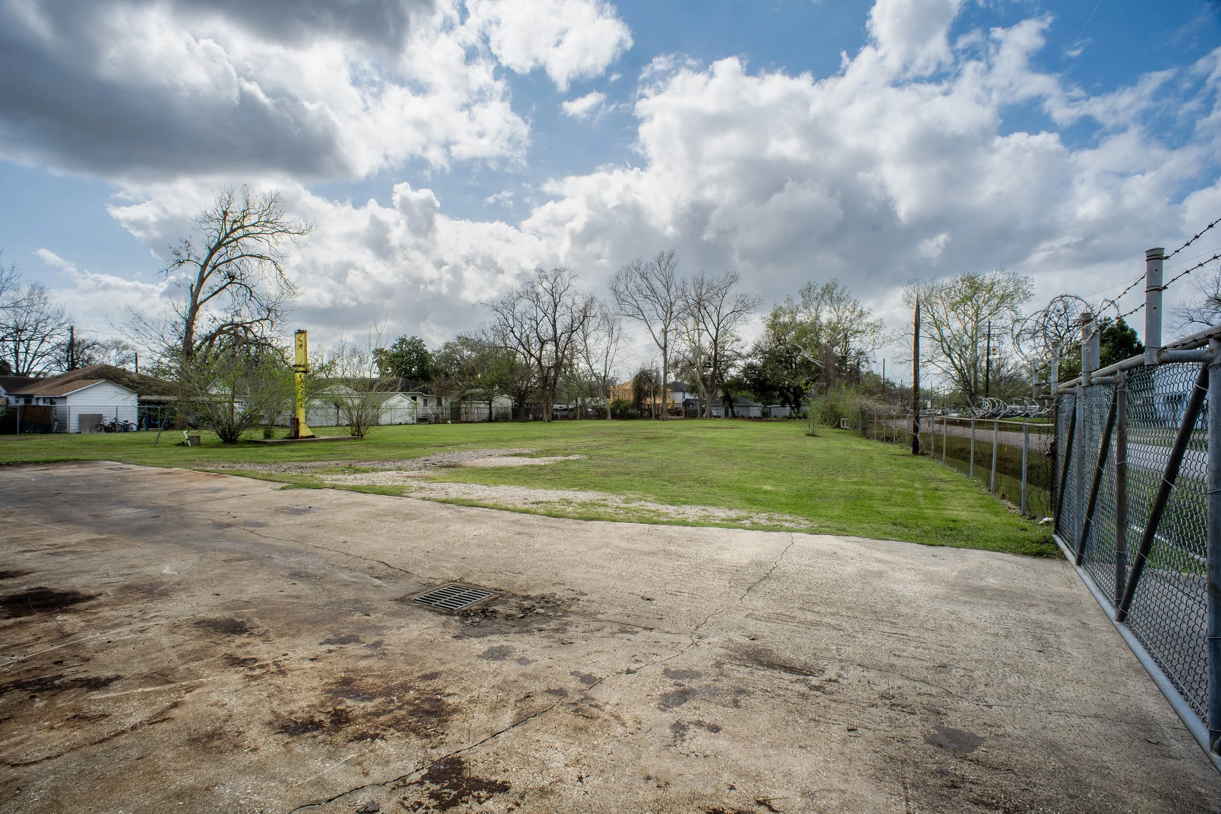 A park with a grassy field, leafless trees, and a partly cloudy sky. There is a chain-link fence on the right side and a weathered concrete area in the foreground.