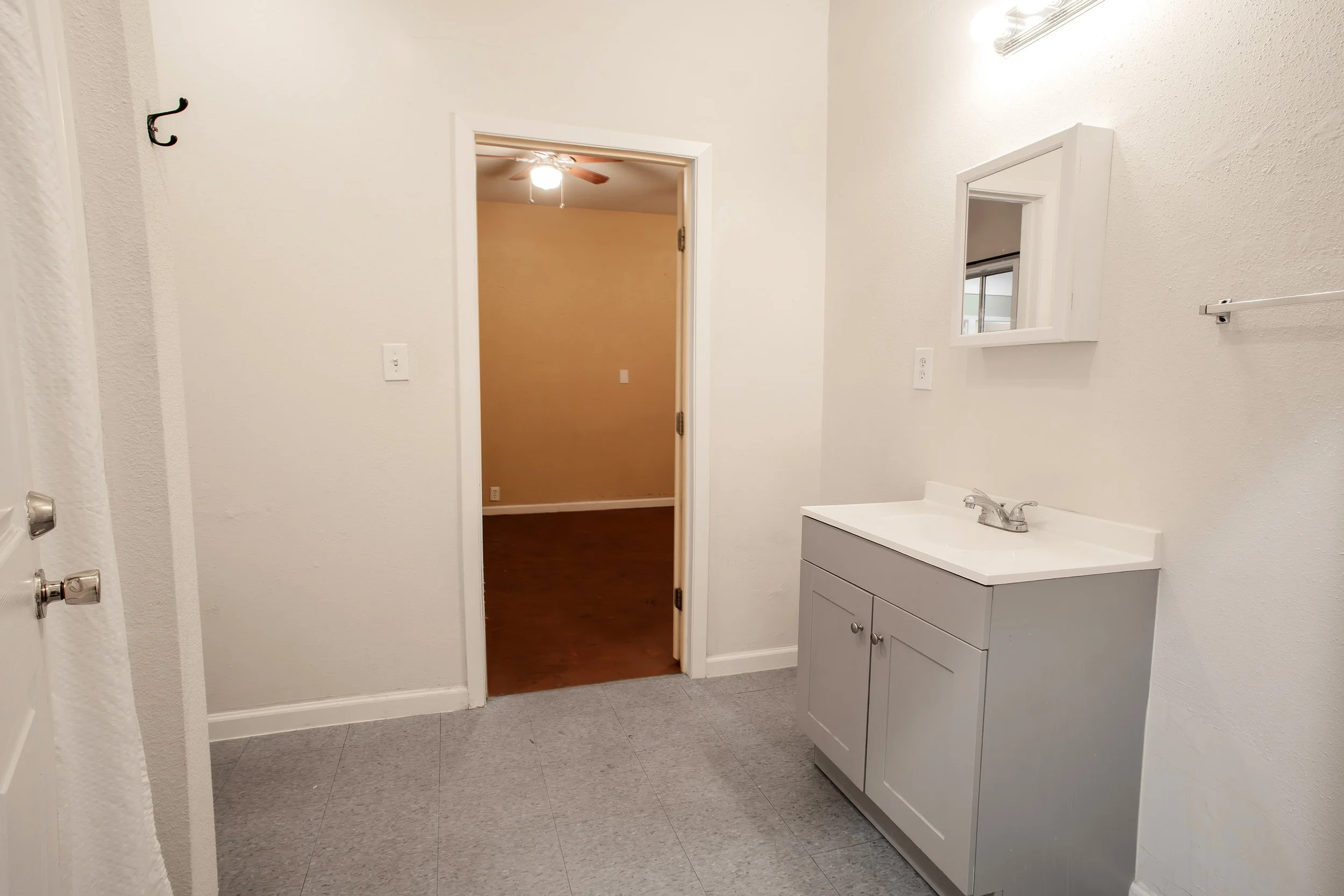 A small bathroom with gray tile flooring, a white vanity with a sink, a wall mirror, a towel rack, and an open doorway leading to a room with a ceiling fan and wooden floor.