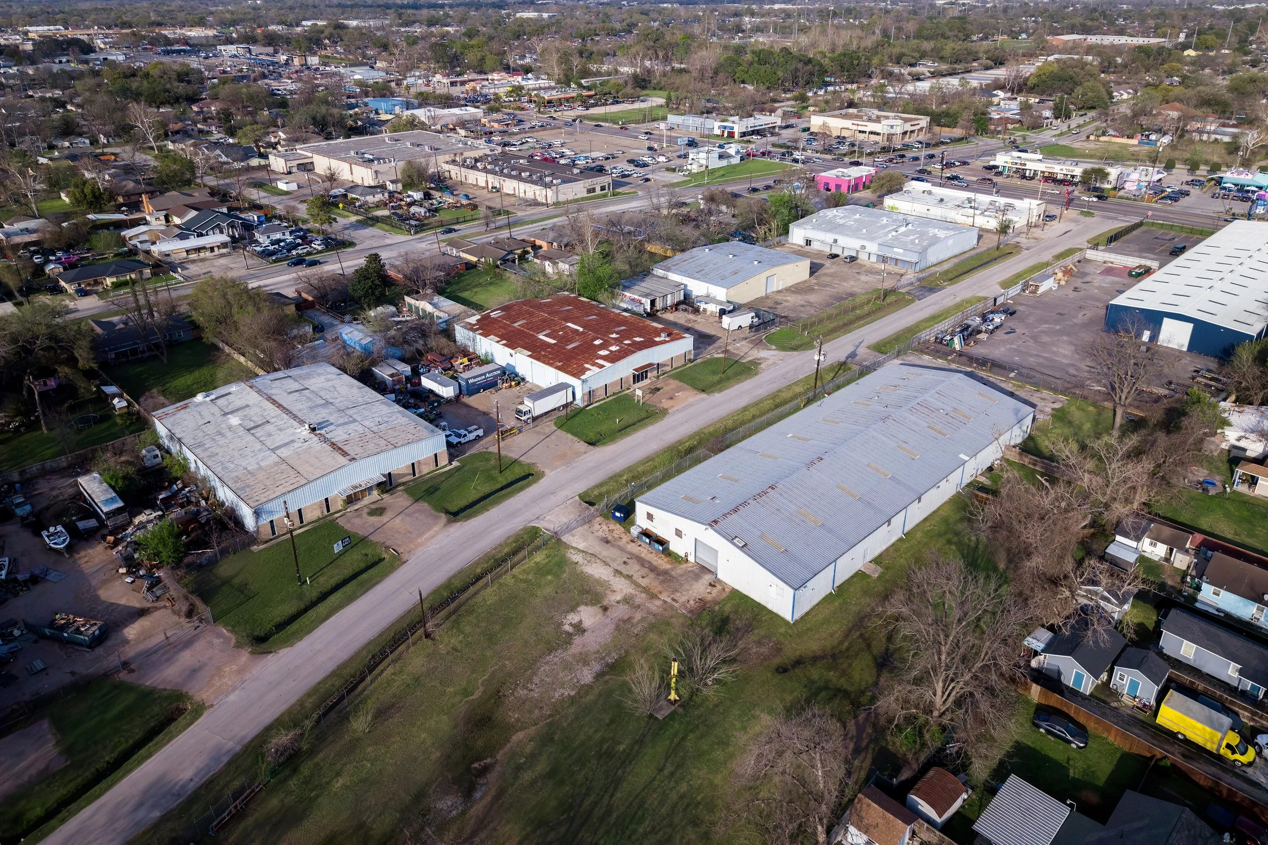 Aerial view of a commercial and residential area with various buildings, parking lots, streets, trees, and a few cars.
