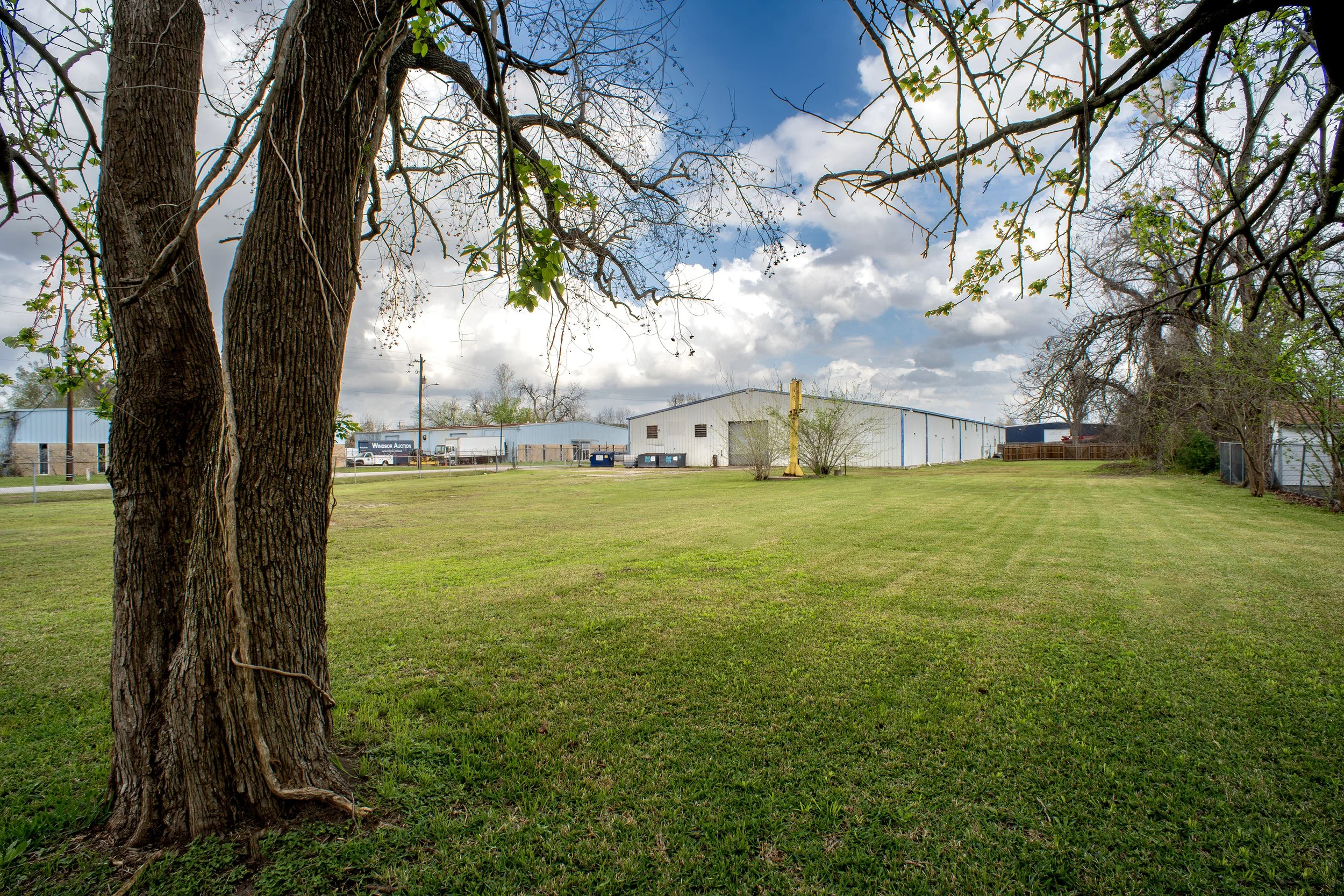 View of a grassy park with large trees in the foreground and industrial buildings in the background under a partly cloudy sky.