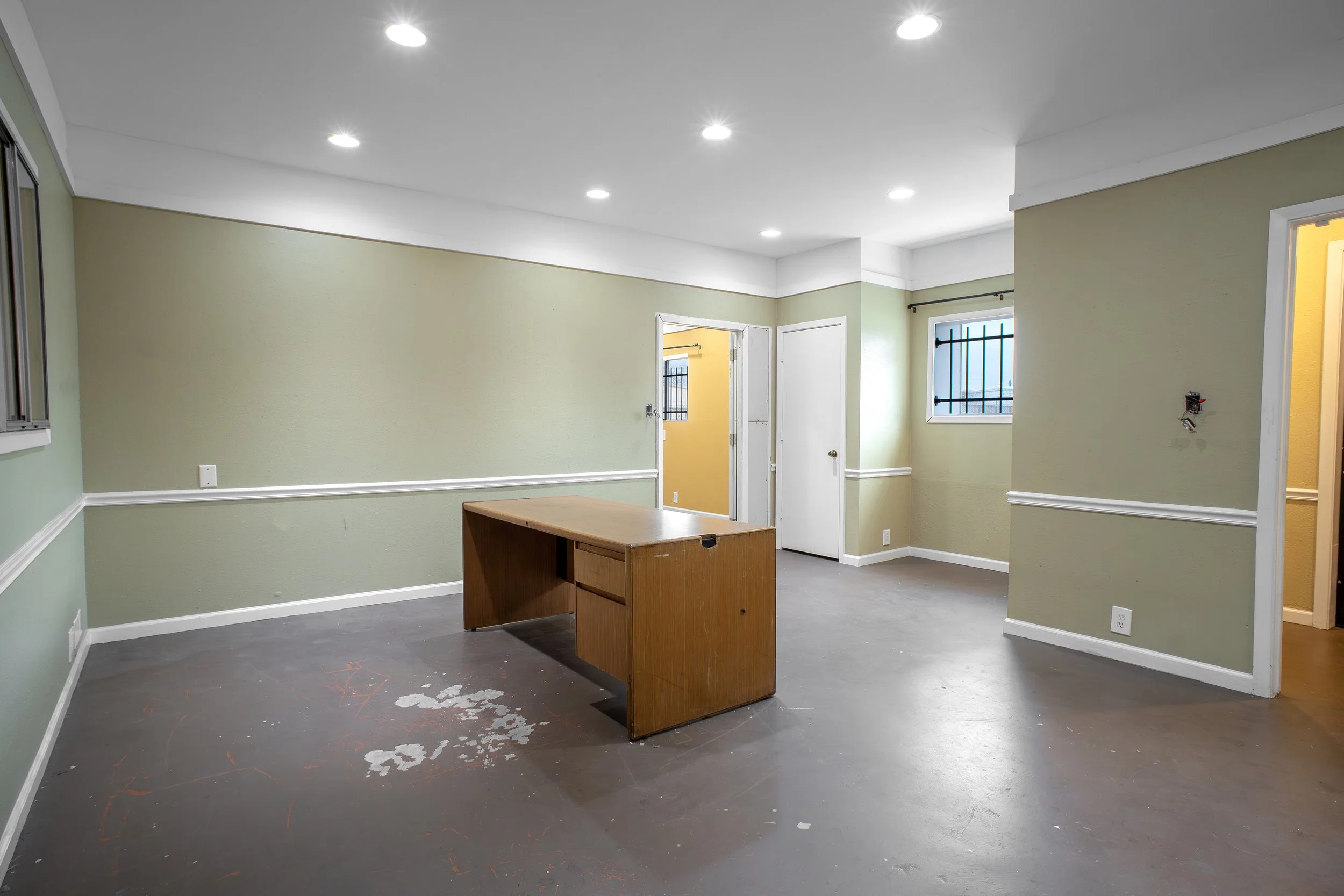An empty room with green walls, a white ceiling with recessed lighting, and a wooden desk on a dark floor with chalk markings. There are doors and windows with barred grilles.