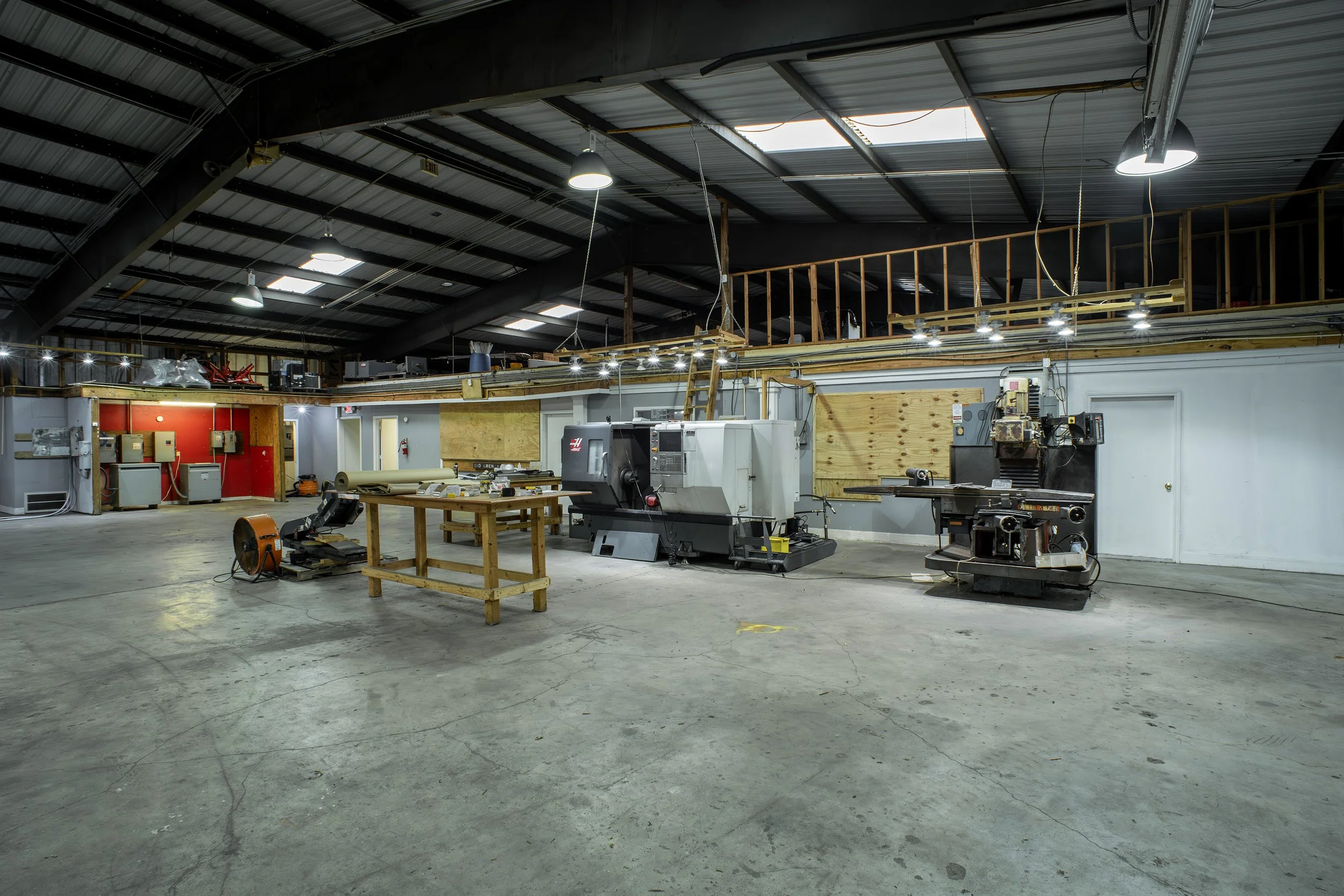 An indoor industrial workspace with high ceilings, ceiling lights, and machinery including a CNC machine, worktables, and tools, with unfinished wooden railings and a second-floor area.
