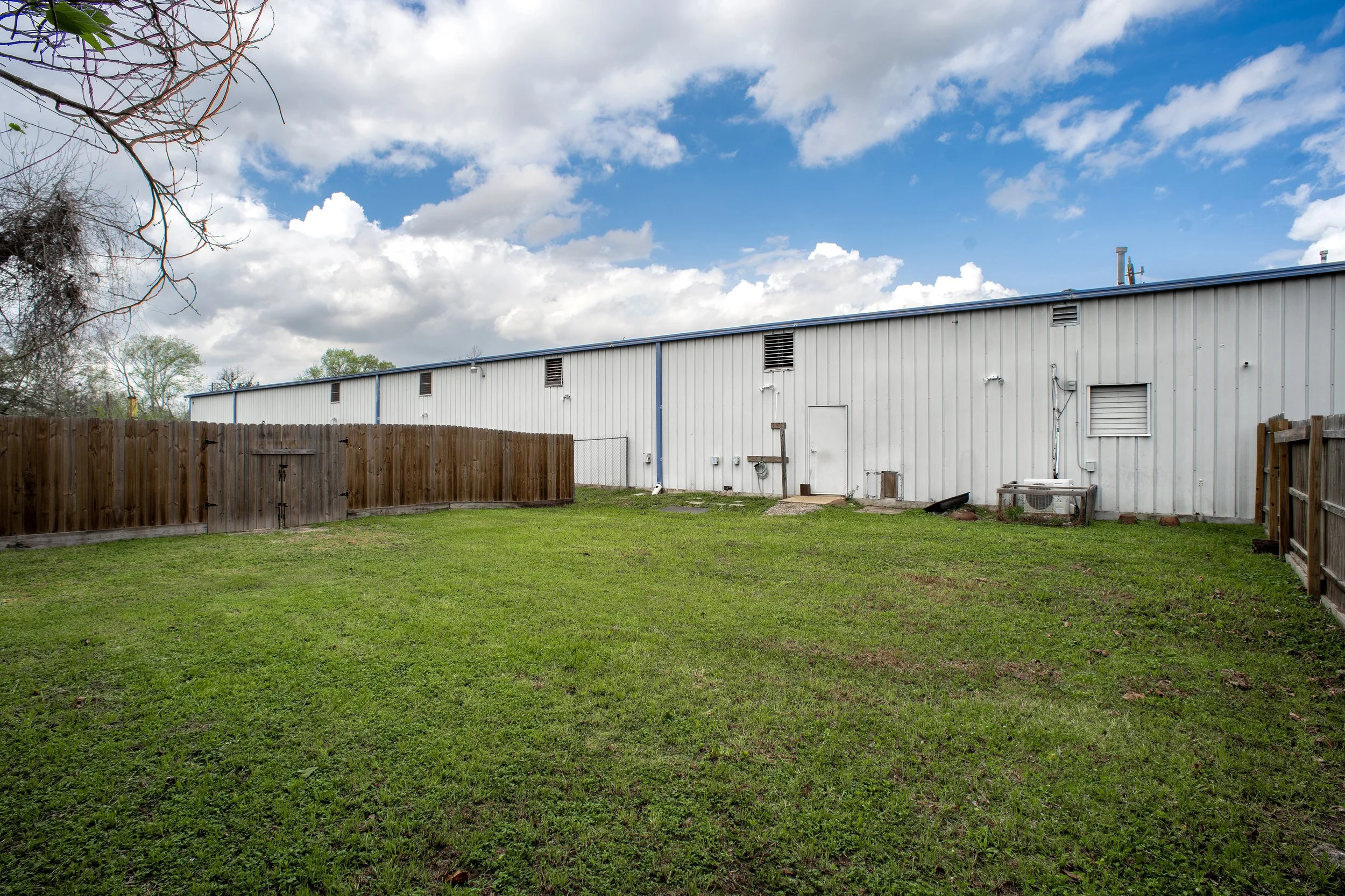 A backyard with a grassy lawn, a wooden fence, and a white industrial building with a metal roof. The sky is partly cloudy with blue patches and white clouds.