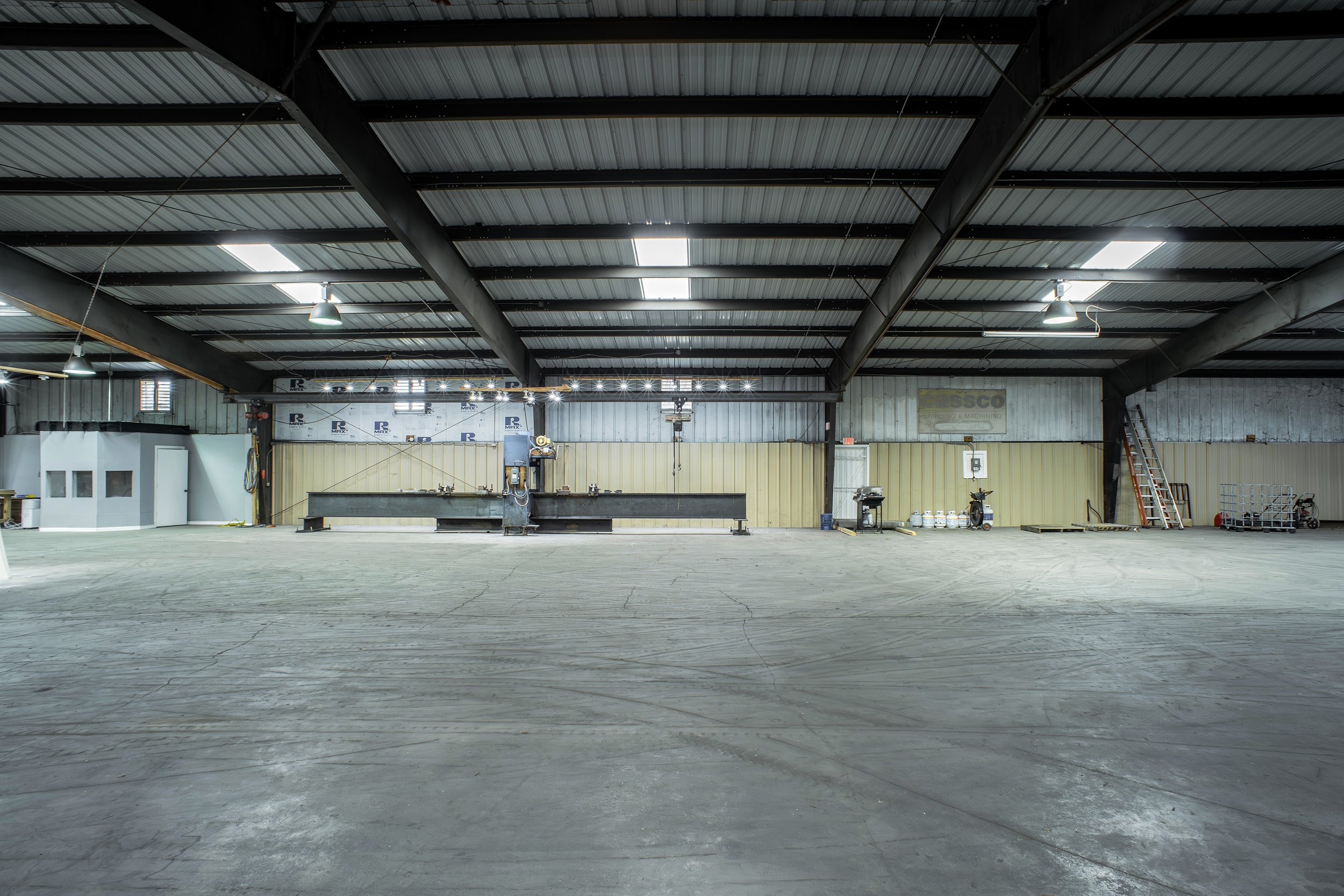 Interior of an empty industrial warehouse with a metal roof, concrete floor, and some equipment and tools along the back wall.