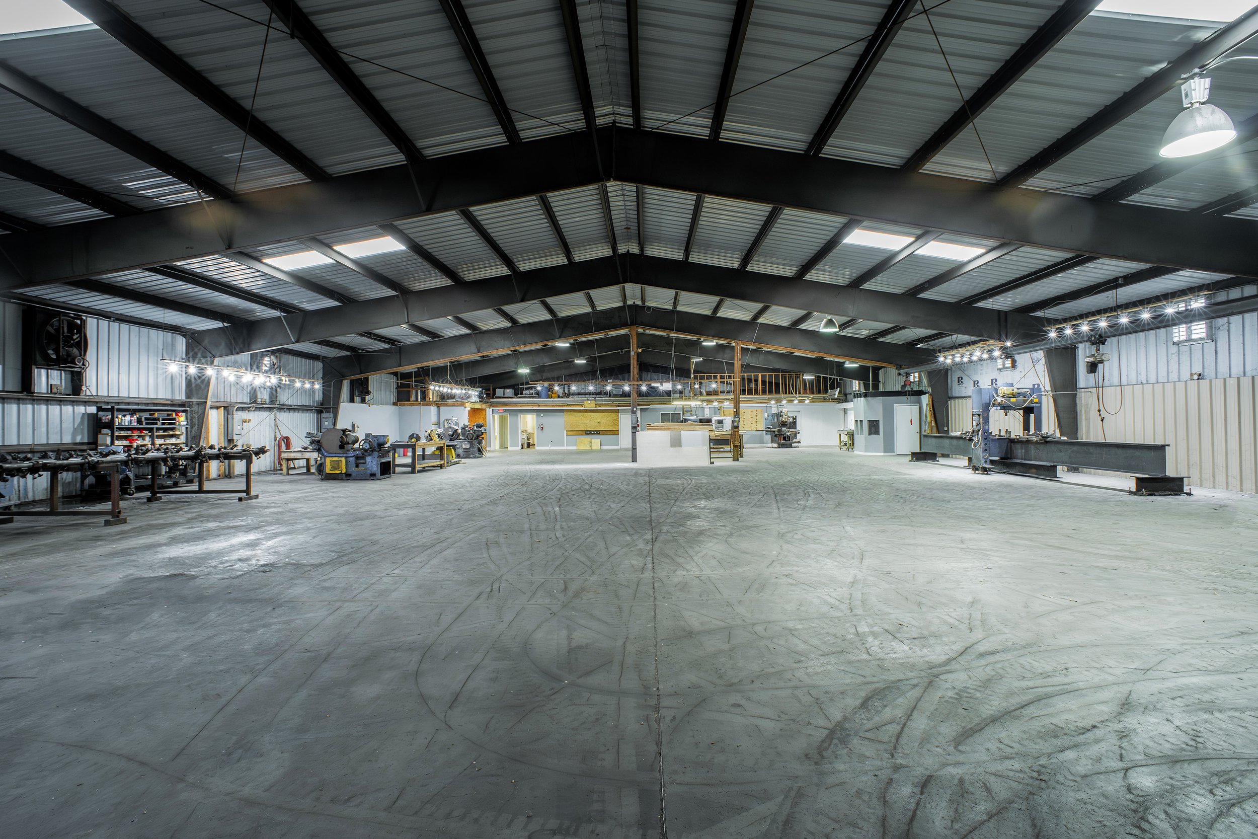 Interior of a large industrial warehouse or workshop with a metal roof, workbenches, machinery, and storage shelves.