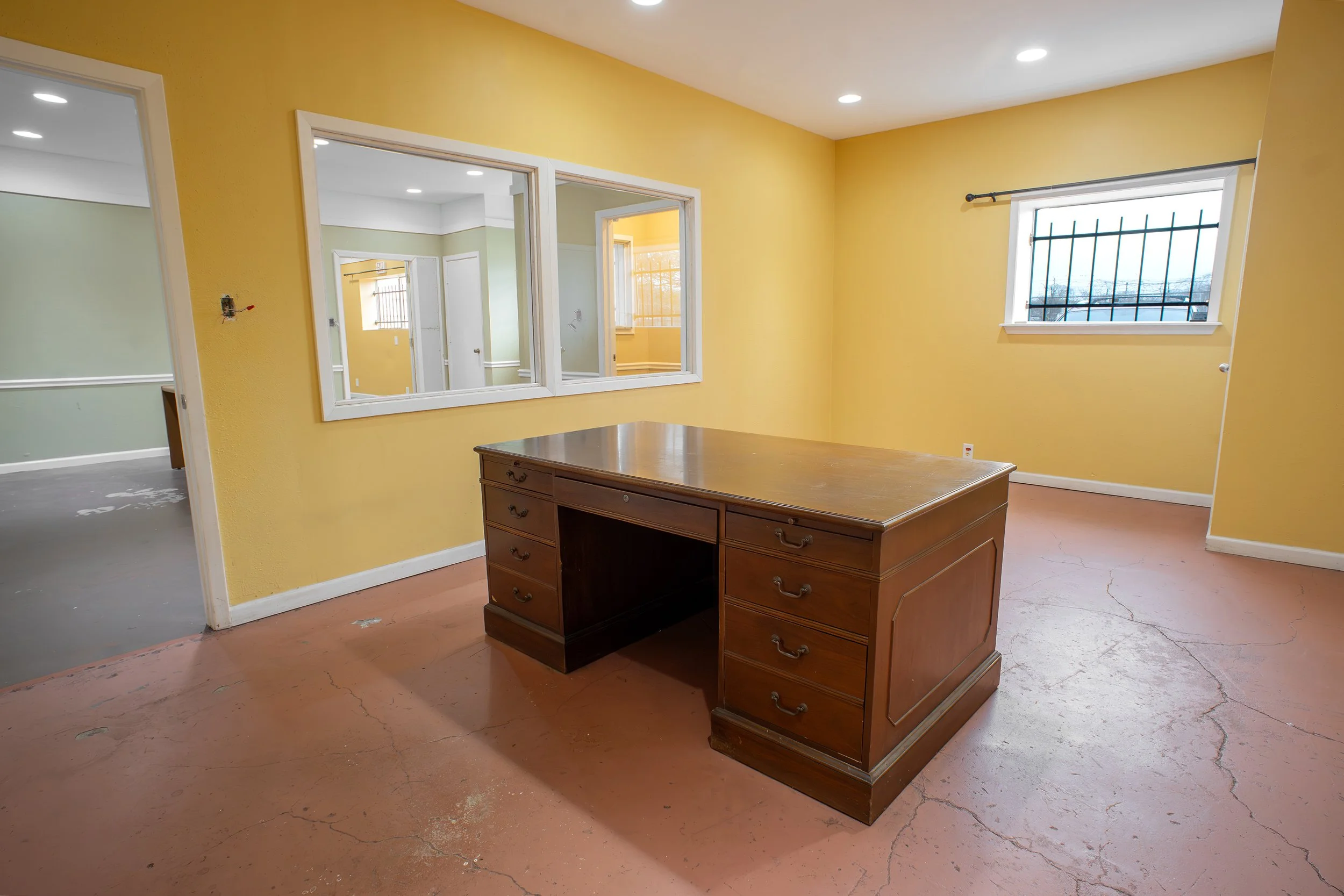 Room with yellow walls, a wooden desk, and a window with bars, showing an outdoor view.