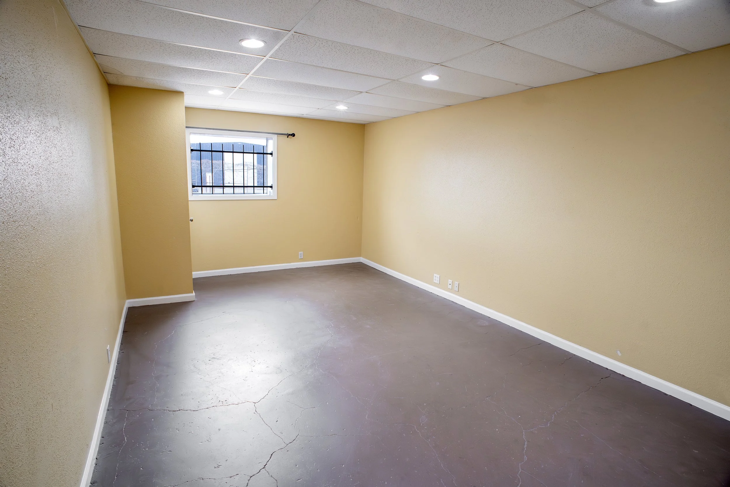 Empty room with yellow walls, a window with bars, a white ceiling with recessed lighting, and a cracked concrete floor.