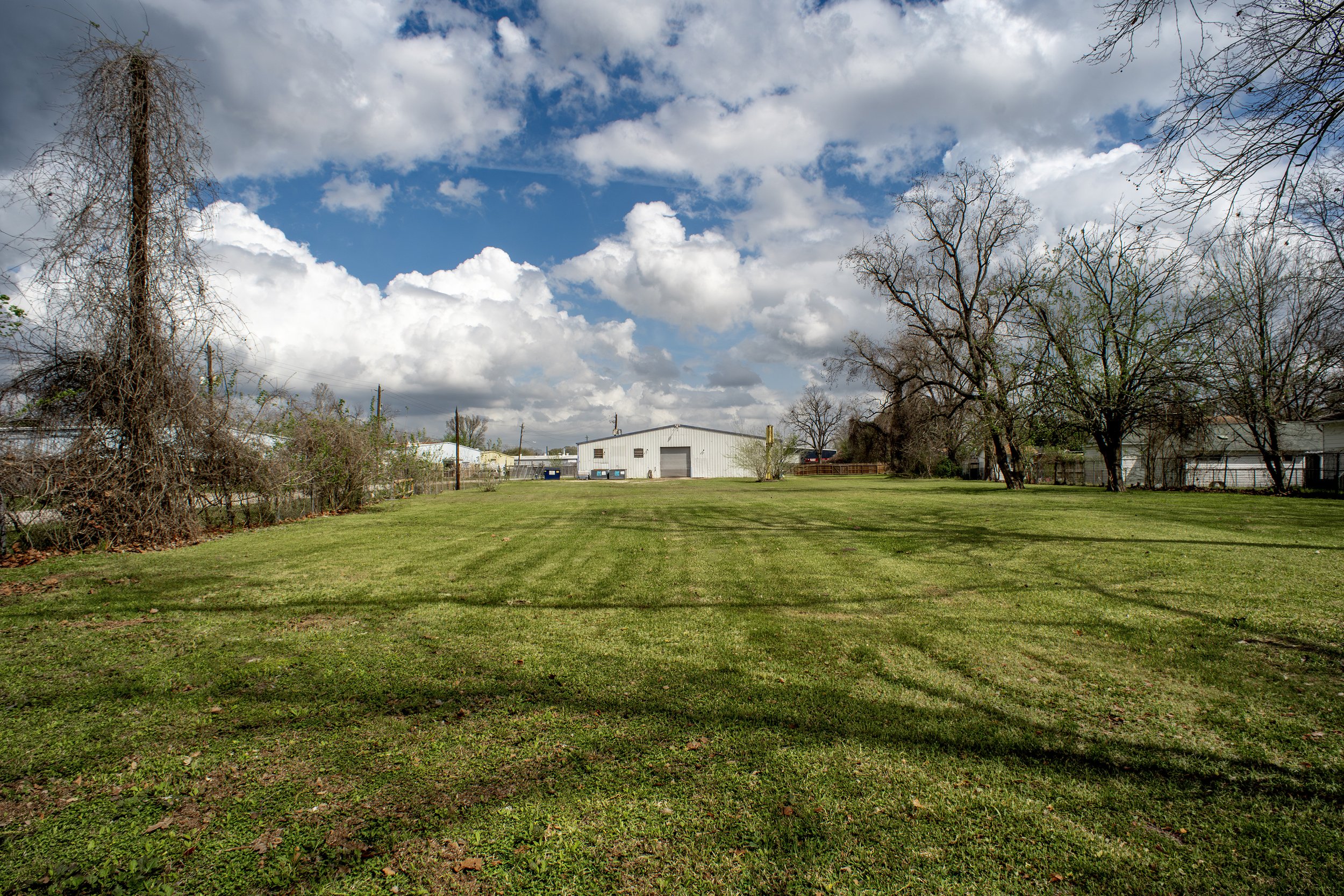 A grassy yard with early spring trees, some without leaves, and a large white metal building in the background under a partly cloudy sky.