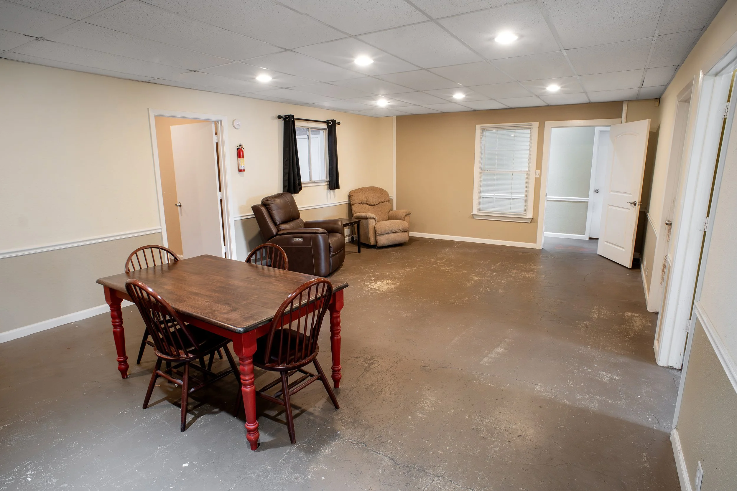 Living room with beige walls, a brown recliner, a beige armchair, a dark wood dining table with red legs, and four wooden chairs. There are two windows with black blinds and curtains, a door on the right, and a fire extinguisher mounted on the wall n