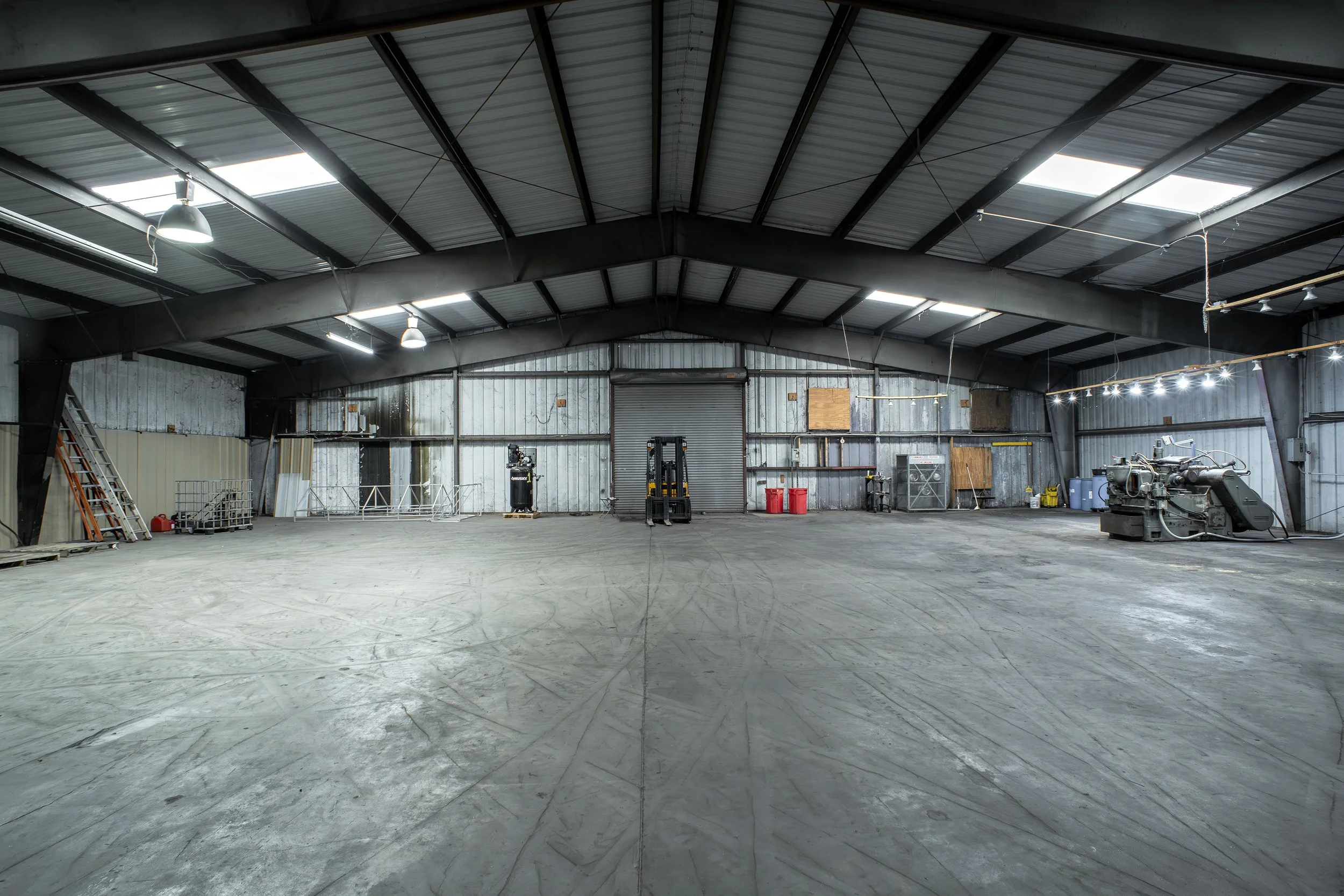 Empty industrial warehouse with metal walls and ceiling, overhead lights, and some equipment and tools along the walls.