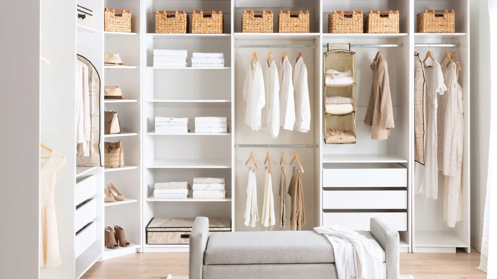 Organized walk-in closet with white shelves and hanging clothes in neutral tones, top baskets, folded linens, shoes, handbags, and a cushioned bench in the foreground.