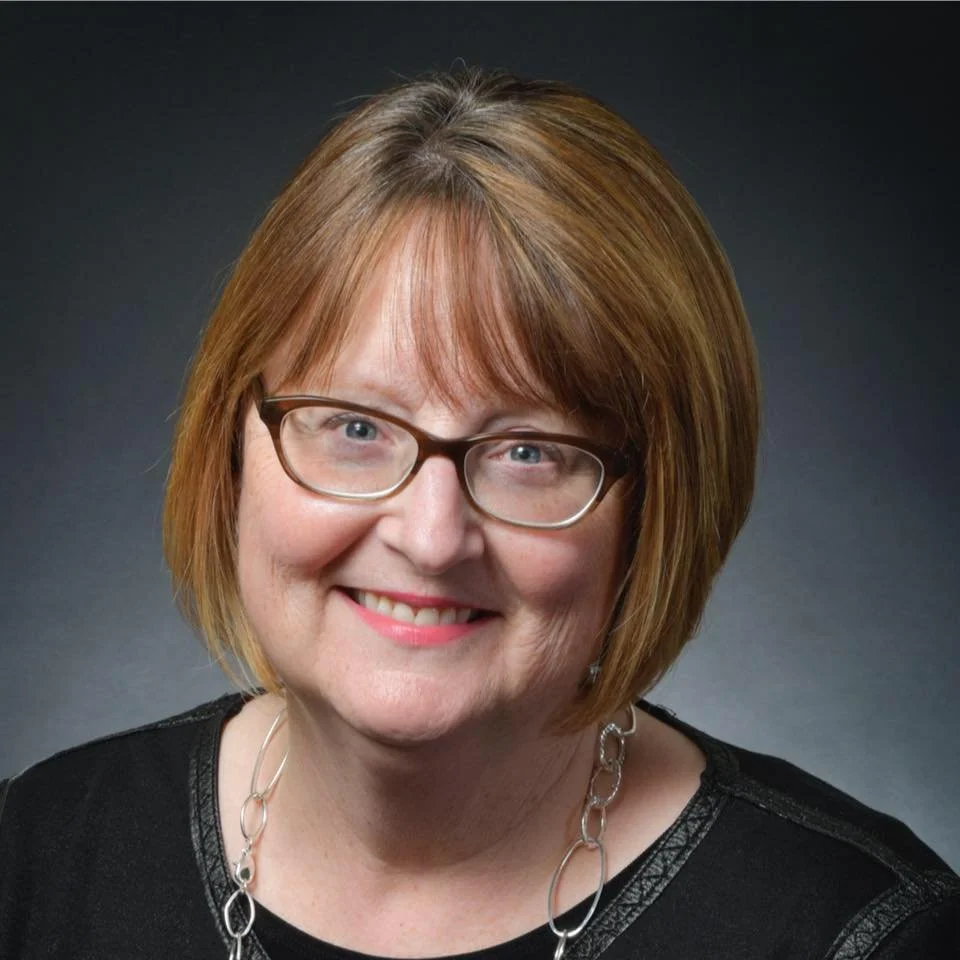 Portrait of a smiling woman with short reddish-brown hair, glasses, wearing a black top and silver necklace against a dark background.