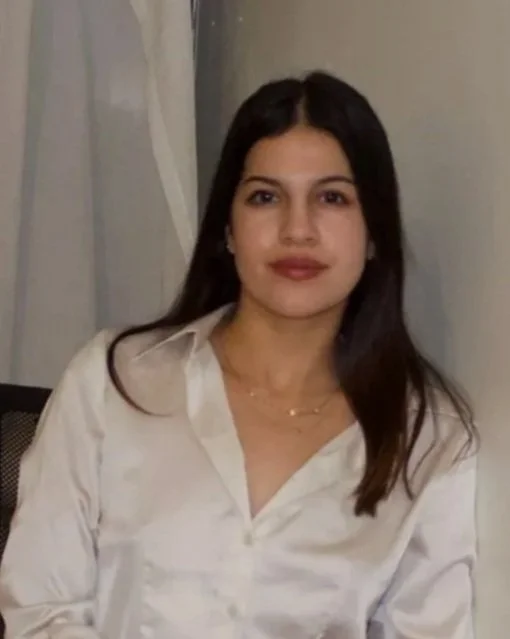 A woman with long dark hair and light makeup sitting indoors, wearing a white button-up shirt.