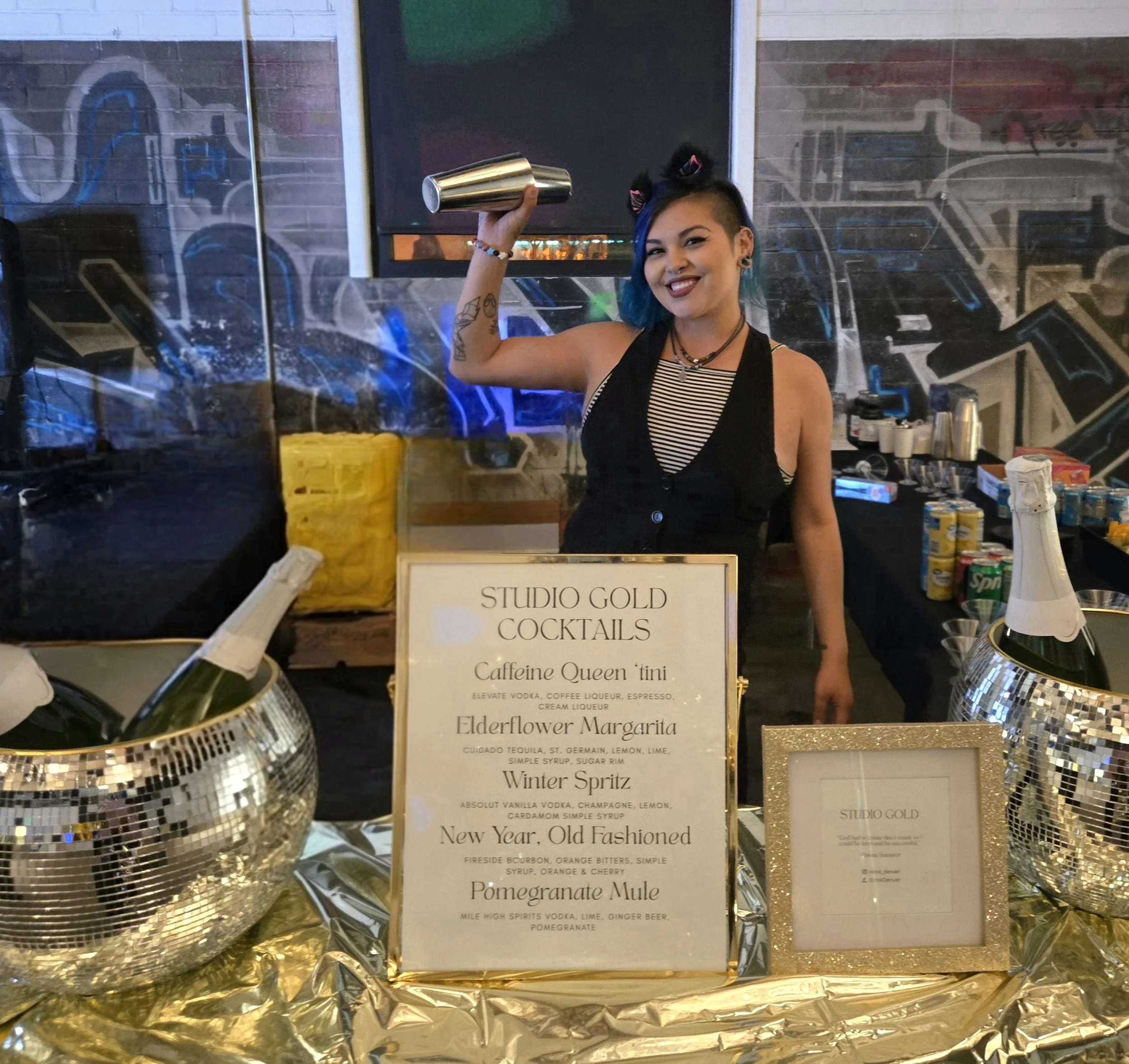 A smiling woman with blue and black hair, wearing a black and white striped top, holding a silver cocktail shaker in a bar setting. Behind her is a menu board listing various cocktails and ingredients, with bottles, cups, and bar tools visible on the counter. The bar area features a reflective, tiled decor with colorful lighting.