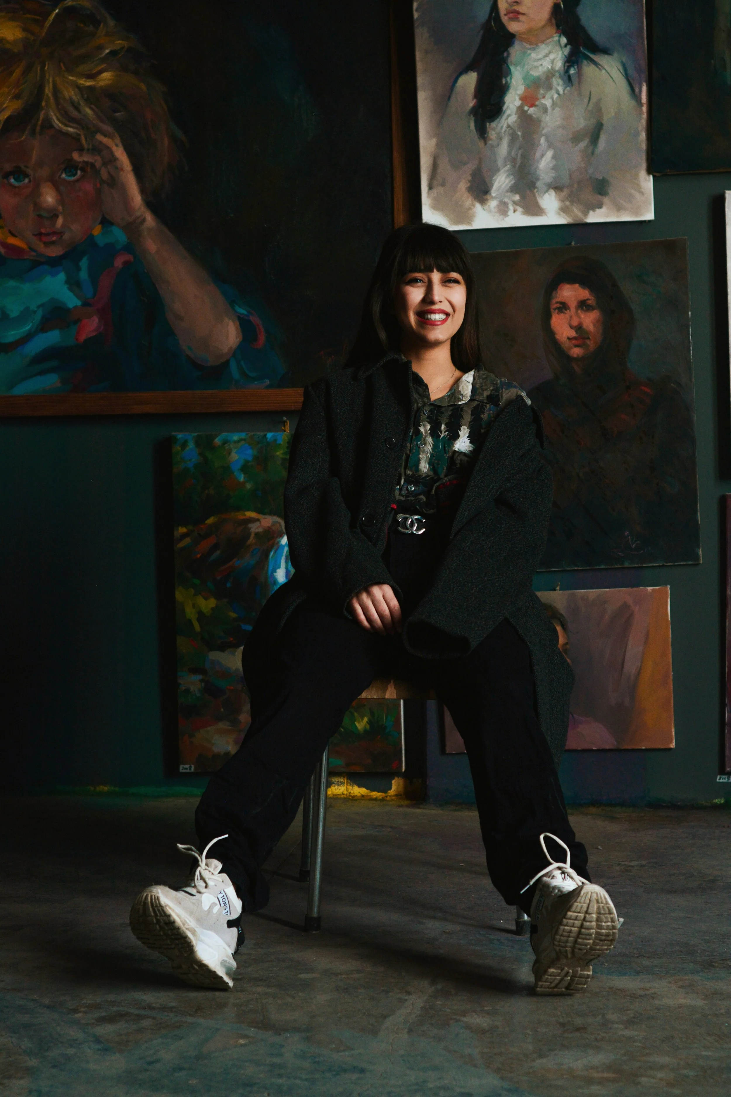 A young woman with dark hair and bangs, wearing a black coat, black pants, and white sneakers, sitting on a chair in an art gallery. She is smiling and holding a black bag. Behind her are several colorful paintings, including portraits and abstract art.